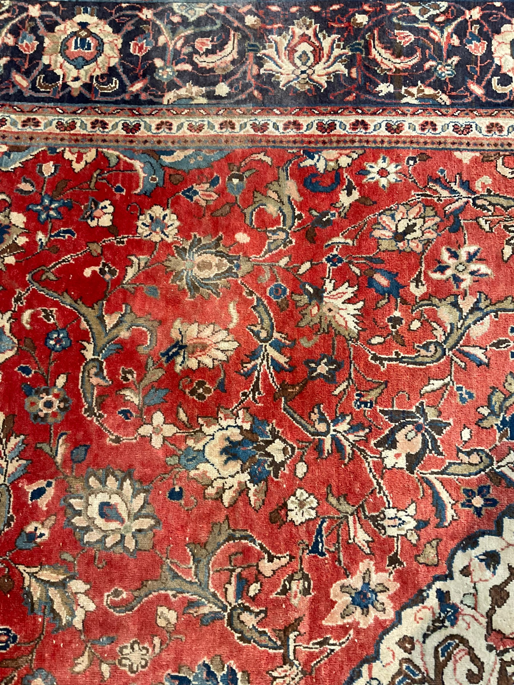 Persian Kashan crimson ground carpet, central shaped pale ground medallion surrounded by interlacing branches and stylised floral motifs, indigo main border with scrolling foliage design, within guard stripes 