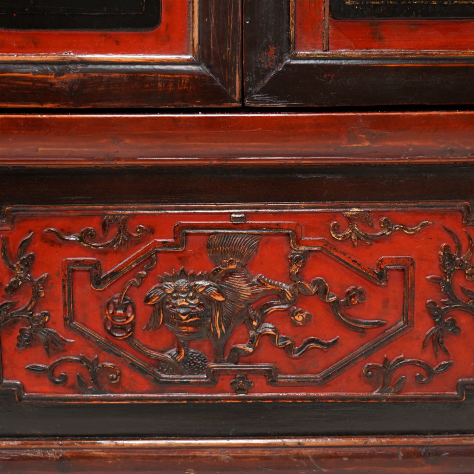 19th century Chinese red and black lacquered hardwood cabinet fitted with four panelled doors, the frieze decorated with scrolling gilt patterns over gold bat mounts surmounting the hinges, the black panels depicting traditional figural and floral scenes, the carved parcel-gilt panels with pagoda, pastoral and war scenes, the doors fitted with metal fish and pagoda handles, enclosing a single shelf and four internal drawers, the base compartment decorated with carved precious objects, raised on moulded square supports with pierced scrolling brackets