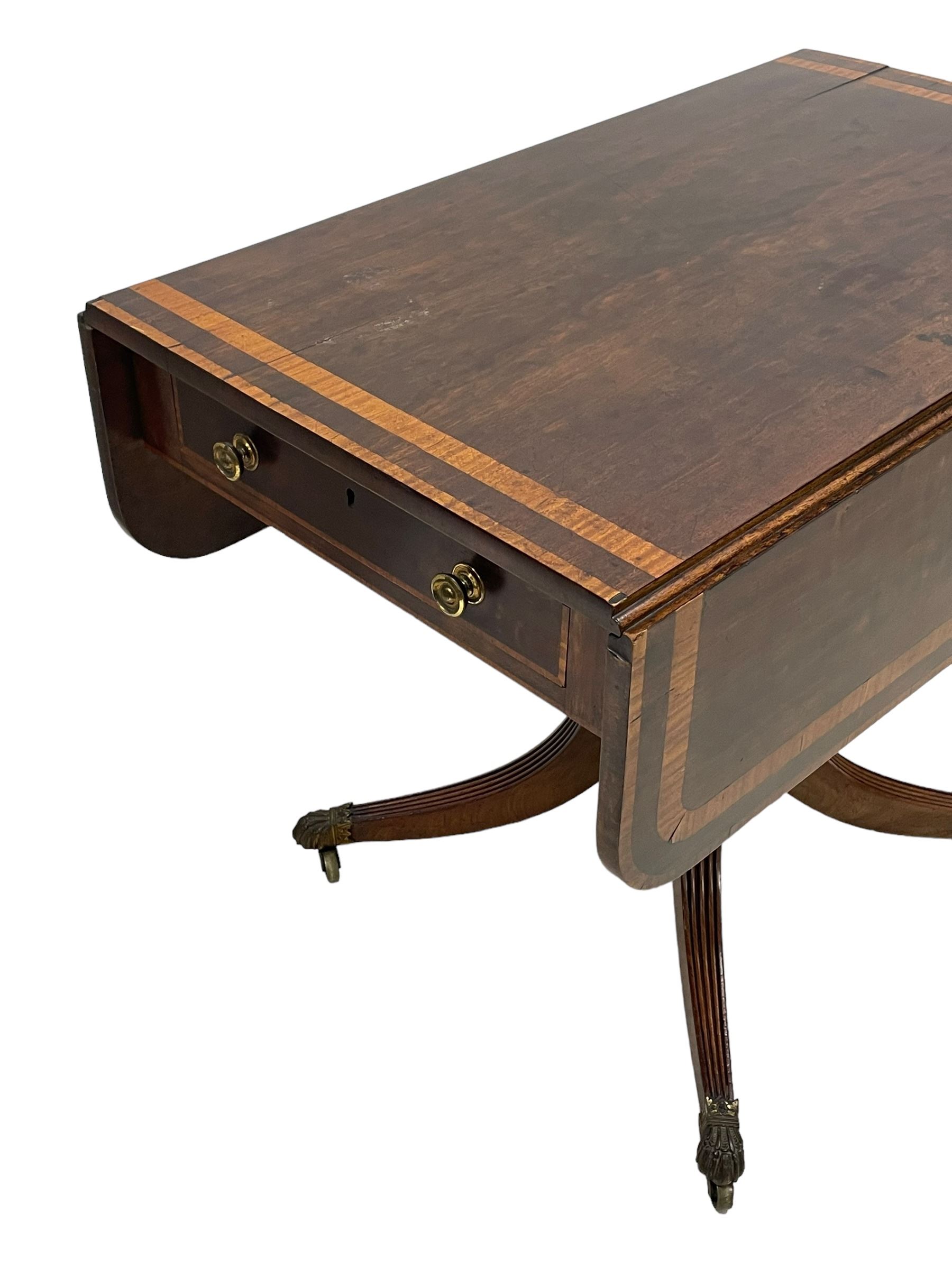 Early 19th century mahogany Pembroke table, rectangular drop-leaf top with rounded corners inlaid satinwood bands, single end drawer and opposing false drawer, turned pedestal on four acanthus carved and reed moulded splayed supports, cast brass cups and castors 