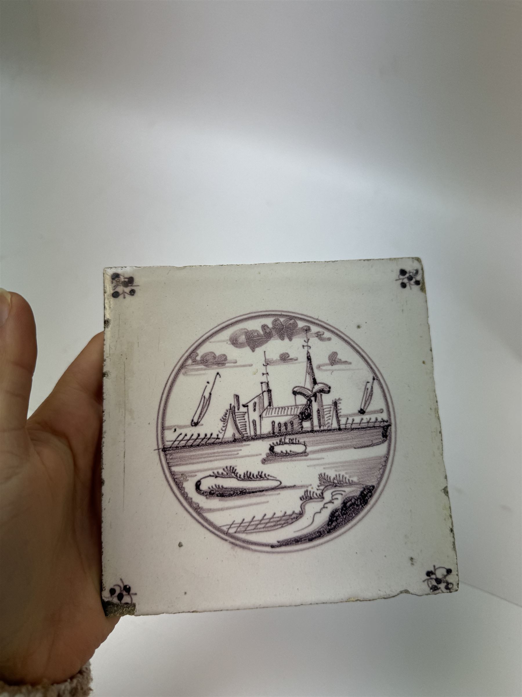 Four Dutch Delft manganese tiles, each depicting a river scene to the centre with a flower to each corner, each tile 12.5cm x 12.5cm