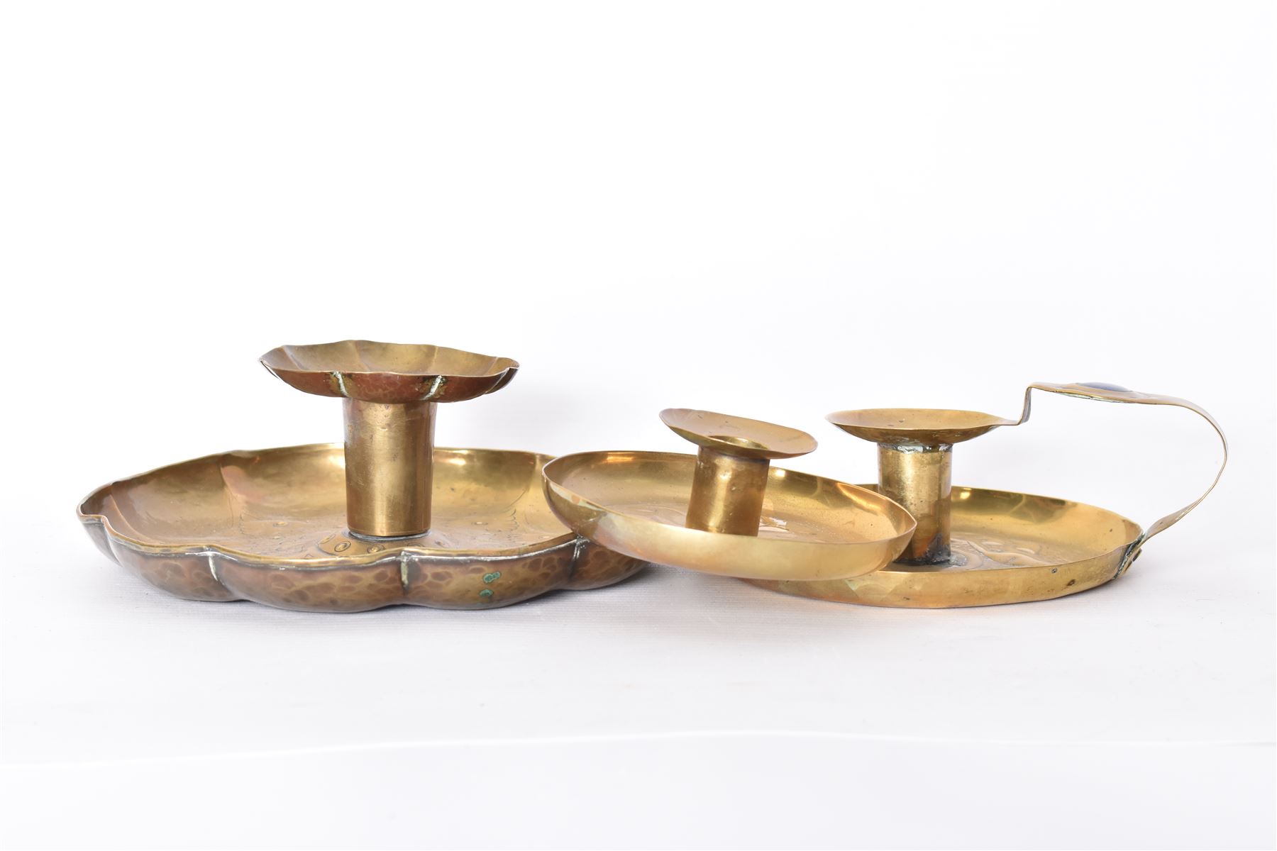 Three Arts & Crafts brass chambersticks/candlesticks, to include a small Keswick School of Industrial Arts example, with repousse leaf decoration, stamped KSIA, a chamberstick with Art Nouveau style decoration to drip pan and blue enamel roundel inset into the loop handle and a larger candlestick, with shaped rim and sconce, repousse decoration and lightly hammered finish, largest D18cm