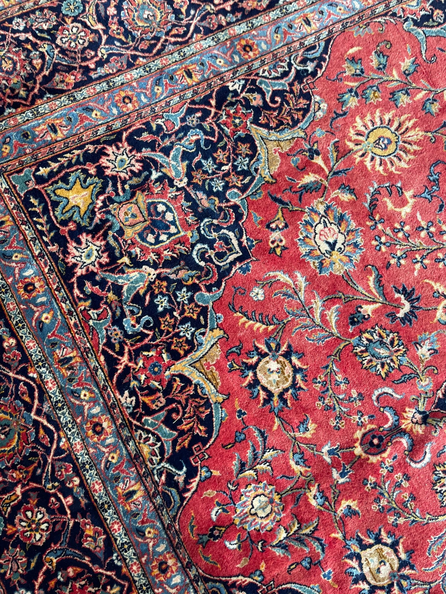 Persian Kashan crimson ground carpet, central rosette medallion on a field of trailing foliate branches decorated with palmettes, scrolling border with repeating plant motifs and flower heads, enclosed within floral pattern guard stripes