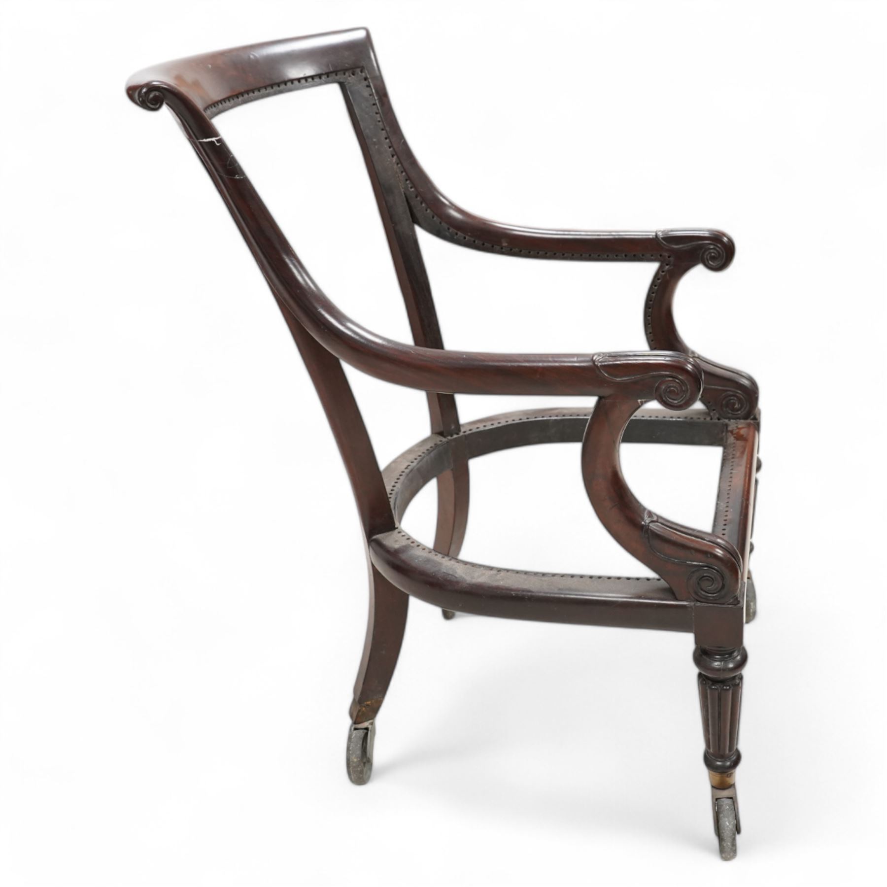 Possibly Gillows - Regency mahogany library chair frame, curved and rolled cresting rail over sweeping arms with lappet and scroll carved terminals, on turned and reed carved supports, brass cups and spoked wheel castors 