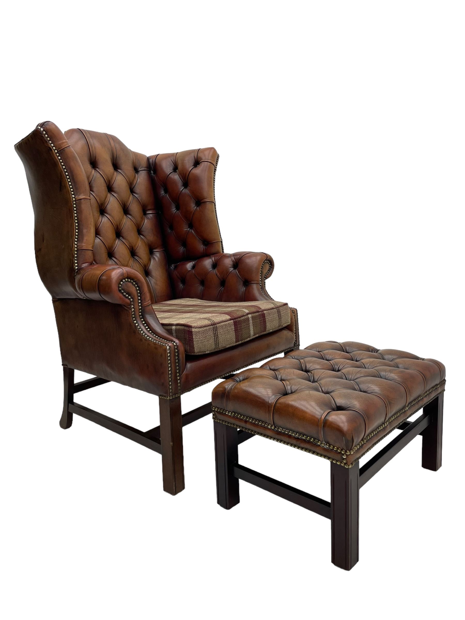 Georgian design hardwood-framed wingback armchair, shaped cresting rail and deep wingback over rolled arms, upholstered in brown buttoned leather with studwork bands, the seat cushion upholstered in checkered fabric, on square supports united by stretchers (W86cm, H118cm, D86cm); together with matching rectangular footstool (63cm x 43cm, H37cm) 