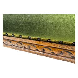 Late Victorian walnut framed sofa, rectangular backrest with carved detailing and central motif, upholstered in olive green fabric with brass studded trim and patterned front rail, scrolled fluted arms supported by turned uprights terminating in bun feet