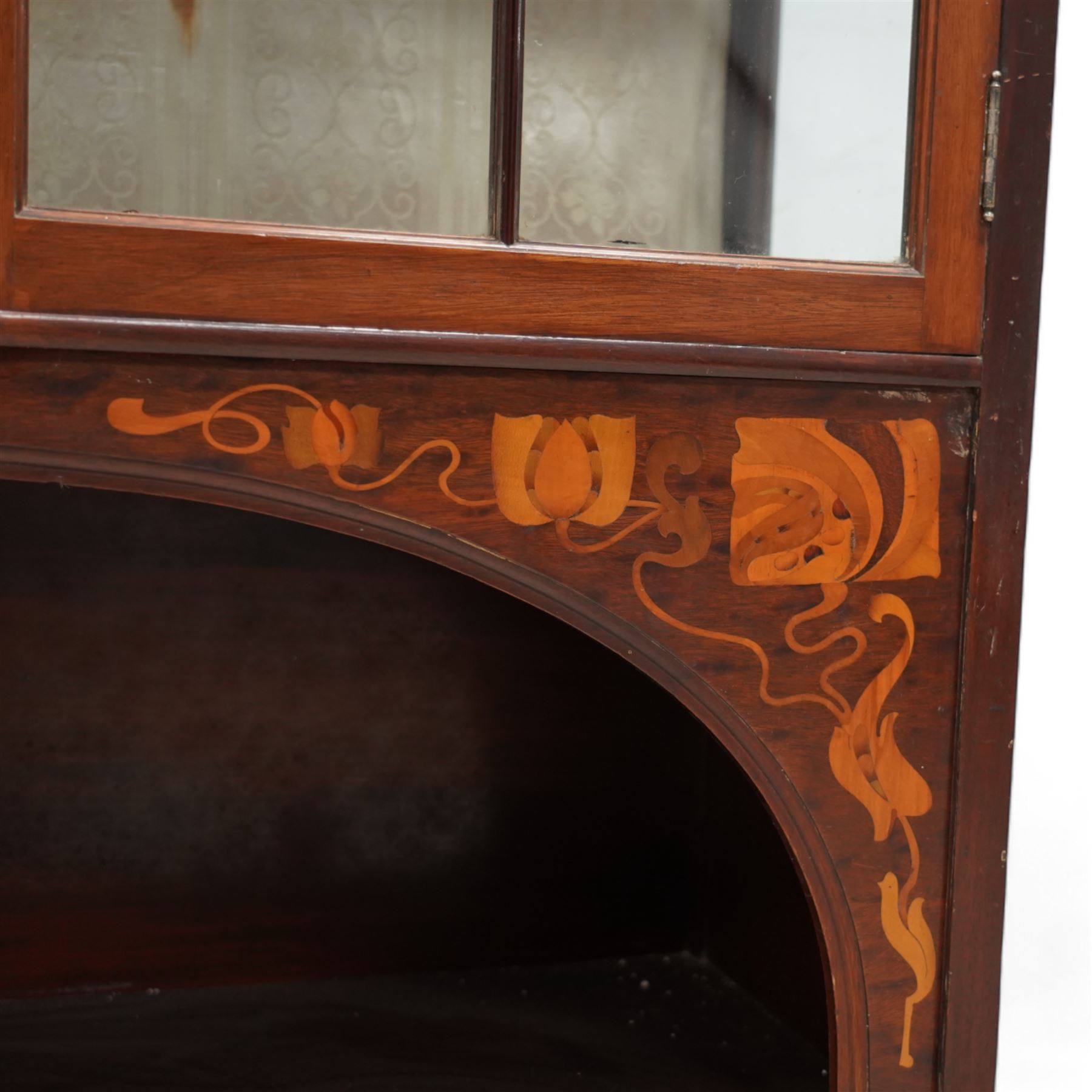 Shapland & Petter; Barnstaple (1854-1999) - Art Nouveau circa. 1900s mahogany display cabinet, raised shelf over panel inlaid with heart and stylised tulips with curled leaves, flanked by balustrade, enclosed by two astragal glazed doors, the lined interior fitted with two shelves, recessed central upright fitted with single shelf, arched lower section fitted with undertier, the spandrels inlaid with further stylised flower heads and trailing branches, on style supports terminating to moulded pad feet, the locks stamped 'S&B, B', retailed by 'Denby & Spinks, Leeds' plaque verso