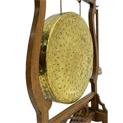 Early 20th century oak gong stand with gong and hammer, shaped cresting rail over moulded upper edge on shaped supports, hanging brass drum gong over fretwork middle rail, on raised platforms terminating to splayed feet