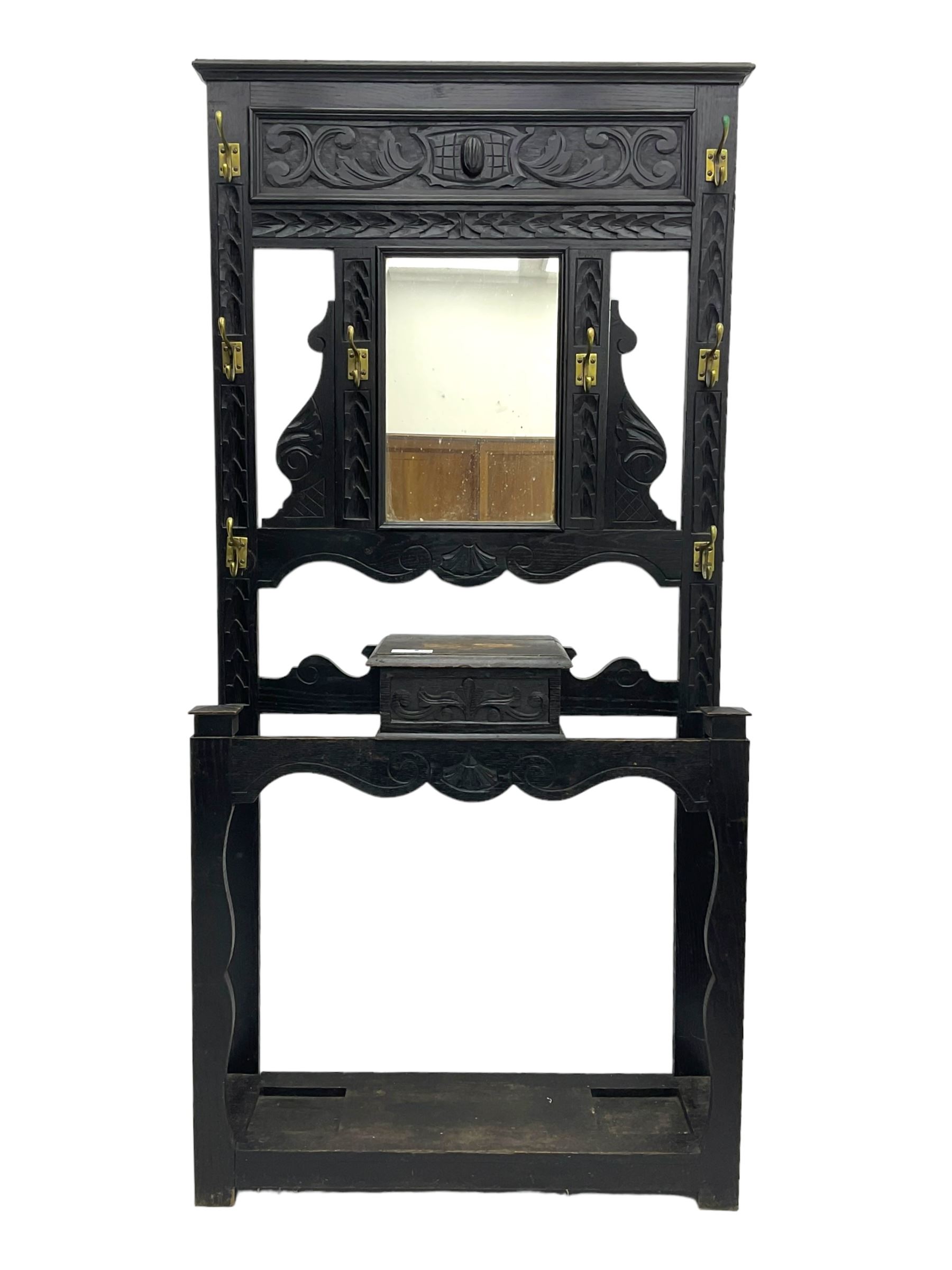 Victorian oak hall stand, the upper section featuring eight brass hooks and central mirror plate flanked by scroll-carved panels, the central section includes a storage box positioned above an umbrella stand, supported by square carved legs with scroll-carved horizontal supports