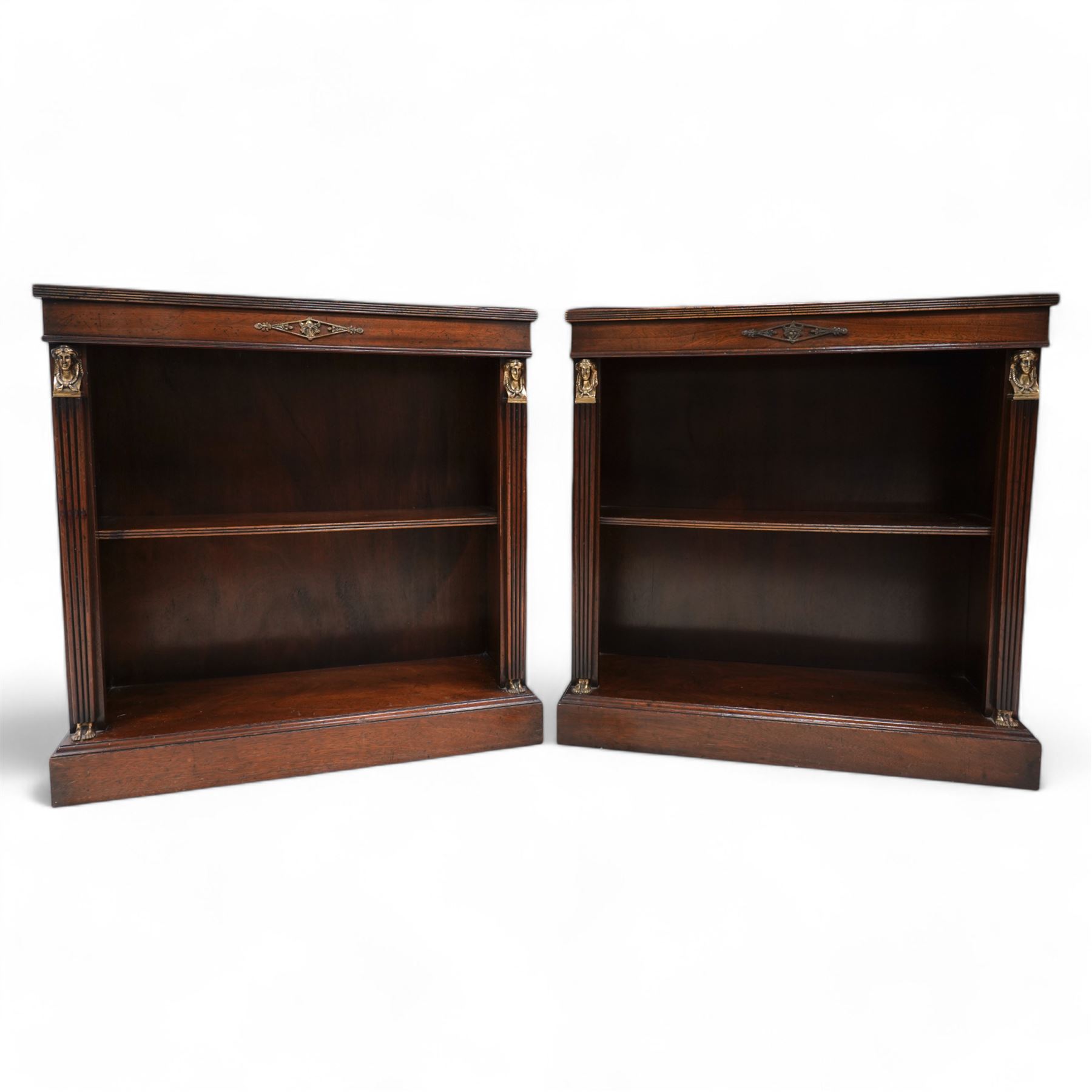 Pair of Neoclassical Empire design mahogany open bookcases, the rectangular top with reeded edge over a plain frieze decorated with a brass mount of the Greek god Hermes, the single shelf flanked by reeded square pilaster uprights, surmounted by a brass capital in the form of a female Egyptian bust, with human feet terminals, lower moulded edge on skirted base