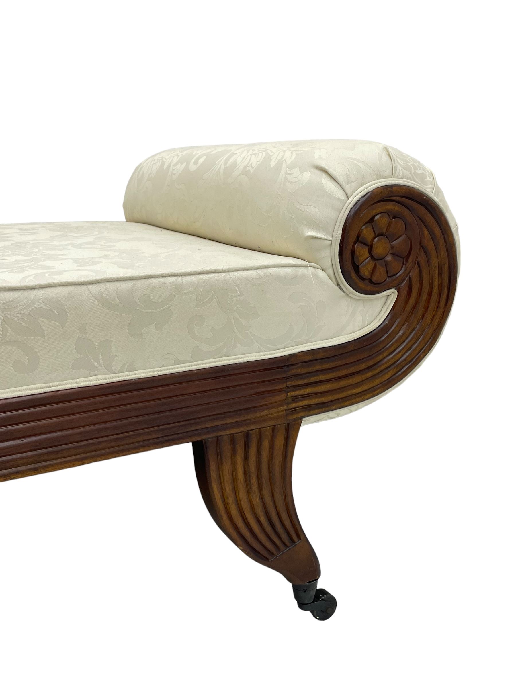 Regency design mahogany chaise longue, scrolled arms with carved rosette details, upholstered in cream damask fabric with bolster cushion, reeded frame supported by turned legs on brass castors