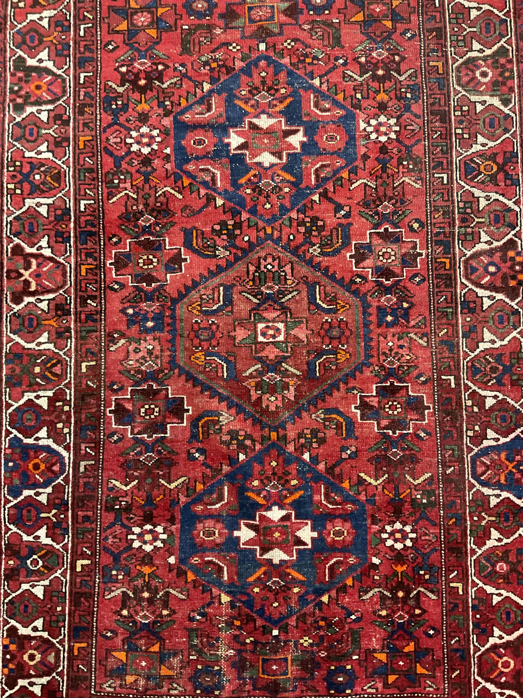 Persian Hamadan rug, red ground with overall geometric design, the field with three extended lozenges, decorated with geometric and stylised flower head motifs, the border and guards decorated with repeating geometric motifs