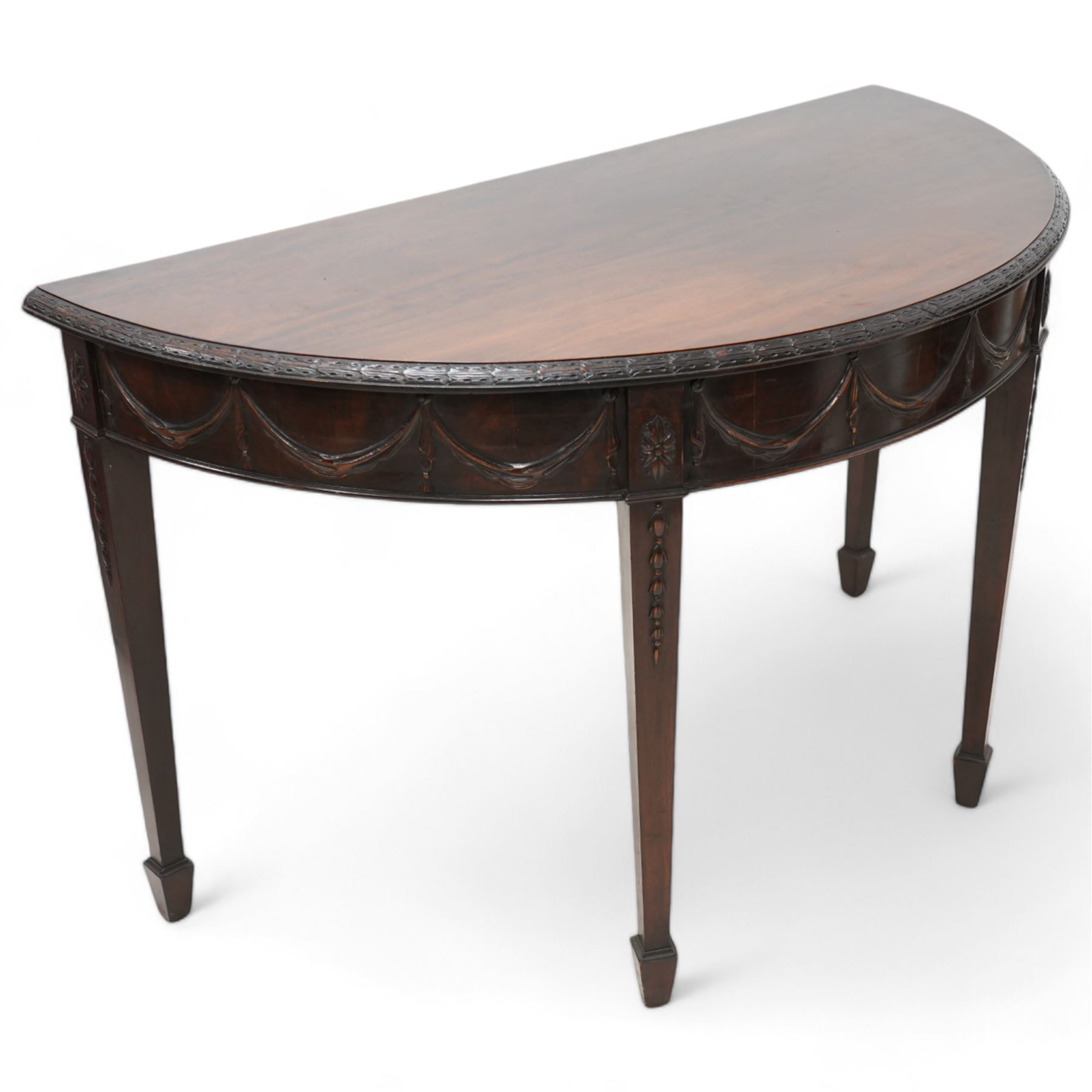 Early 20th century Hepplewhite Revival mahogany console table, demi-lune top with carved moulded edge, the frieze decorated with moulded repeating festoons, raised on square tapering supports with applied bell-flower motifs, terminating in spade feet