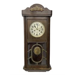 Oak cased 1930s 8-day German wall clock - with a crested top, glazed door and visible pendulum, silvered dial with Arabic numerals and spade hands, two train movement striking the hours on a gong. With pendulum and key.