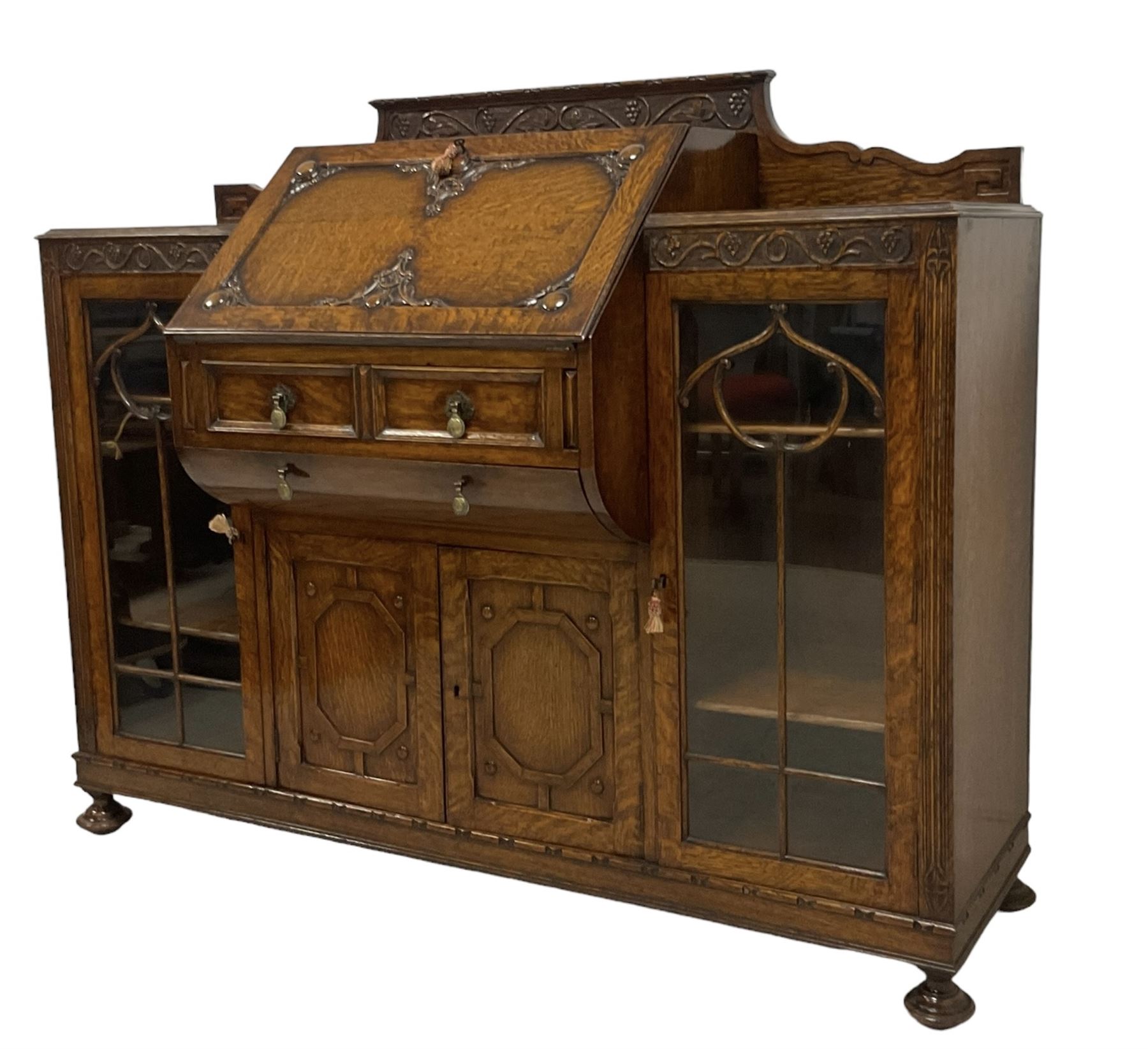 Early 20th century heavily carved oak bookcase bureau, raised back with grape-vine carved frieze, over fall-front enclosing fitted interior, above two short drawers and curved frieze drawer, flanked by astragal glazed and panelled cupboards, on turned feet