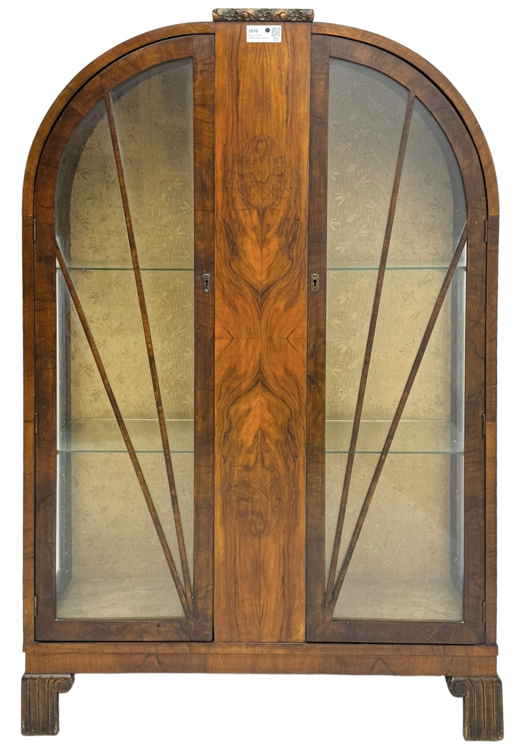 Early 20th century Art Deco walnut display cabinet, with arched top and carved floral crest, central band flanked by two glazed panel doors with decorative mullions, enclosing two glass shelves, on scroll carved bracket feet