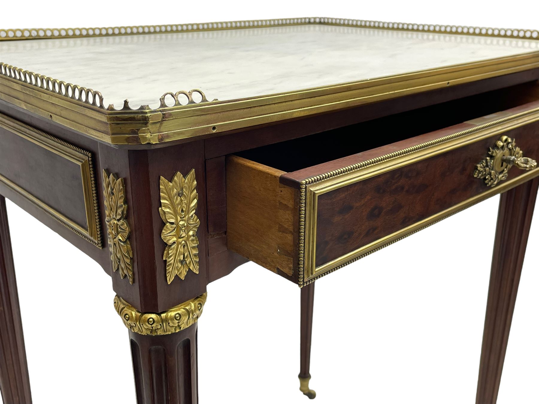 Late 19th century French figured plum pudding mahogany side table, canted rectangular white marble top with raised gilt metal gallery, fitted with a single frieze drawer, central extending floral cast ormolu escutcheon and applied moulded edging, on tapering turned and fluted supports with brass cups and castors, decorated with cast foliate mounts and foliate cast collars 