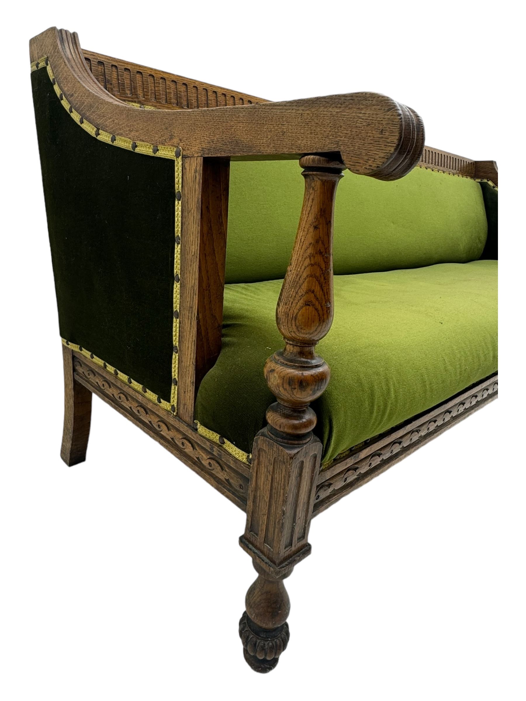 Late Victorian walnut framed sofa, rectangular backrest with carved detailing and central motif, upholstered in olive green fabric with brass studded trim and patterned front rail, scrolled fluted arms supported by turned uprights terminating in bun feet