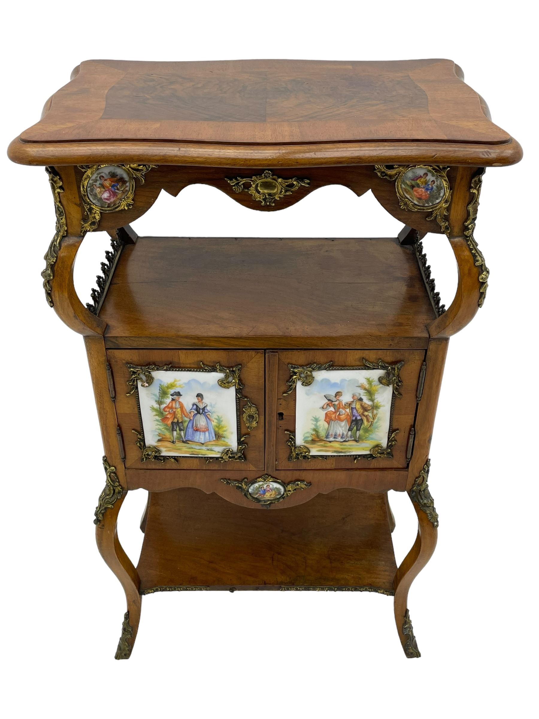 Mid-to-late 20th century French figured walnut three-tier bedside stand, shaped moulded top inlaid with figured book-matched veneers and crossbanding, double cupboard below enclosed by two doors mounted by painted porcelain panels depicting courting scenes within landscapes, on cabriole supports united by undertier, decorated with ornate cast metal cartouche mounts 