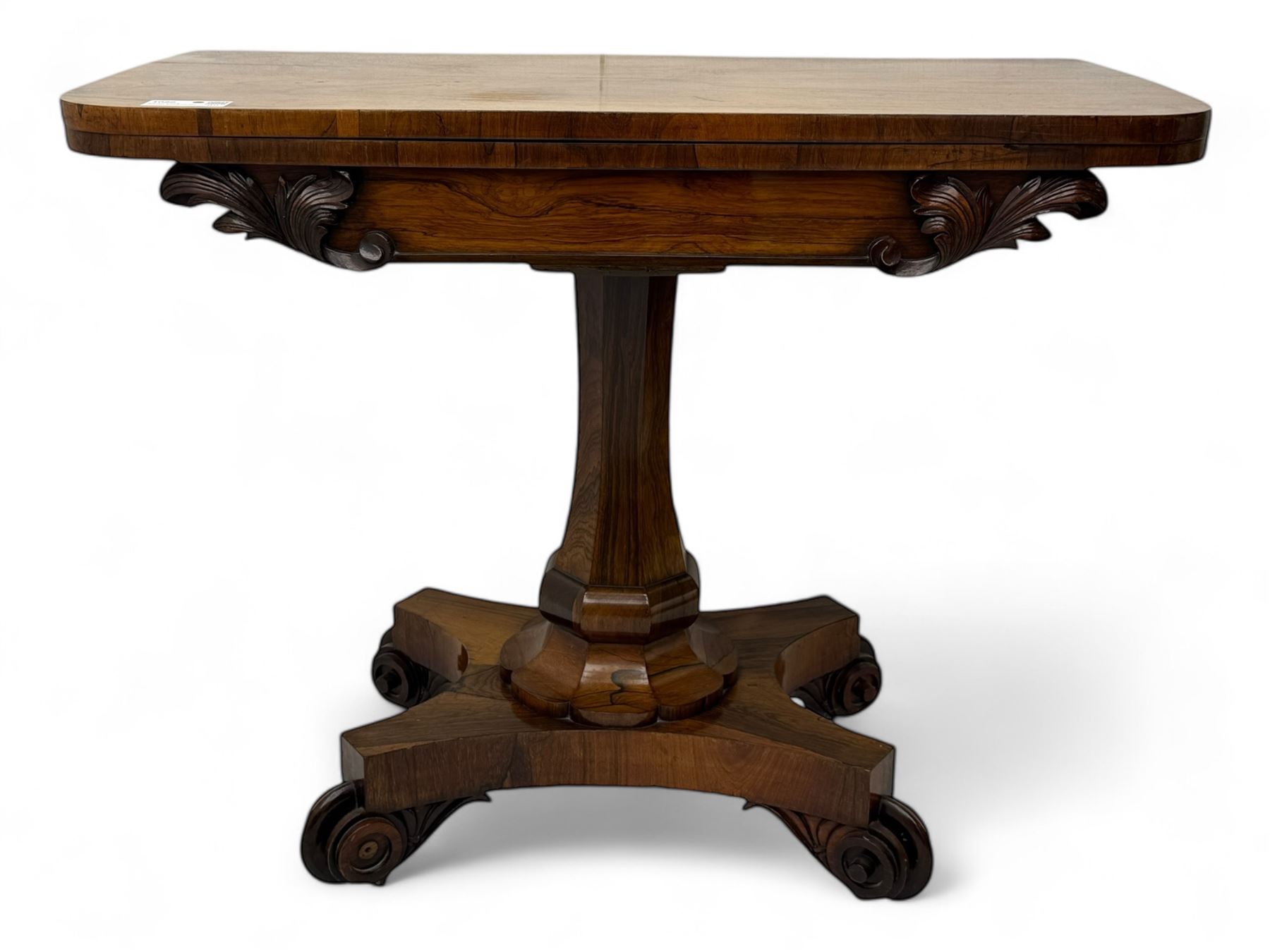 Early Victorian rosewood card table, rectangular foldover swivel top with rounded corners, red baize-lined surface, frieze adorned with carved foliate motifs, supported by a central octagonal waisted column on shaped quatrefoil base, terminating in scrolled feet with concealed metal castors