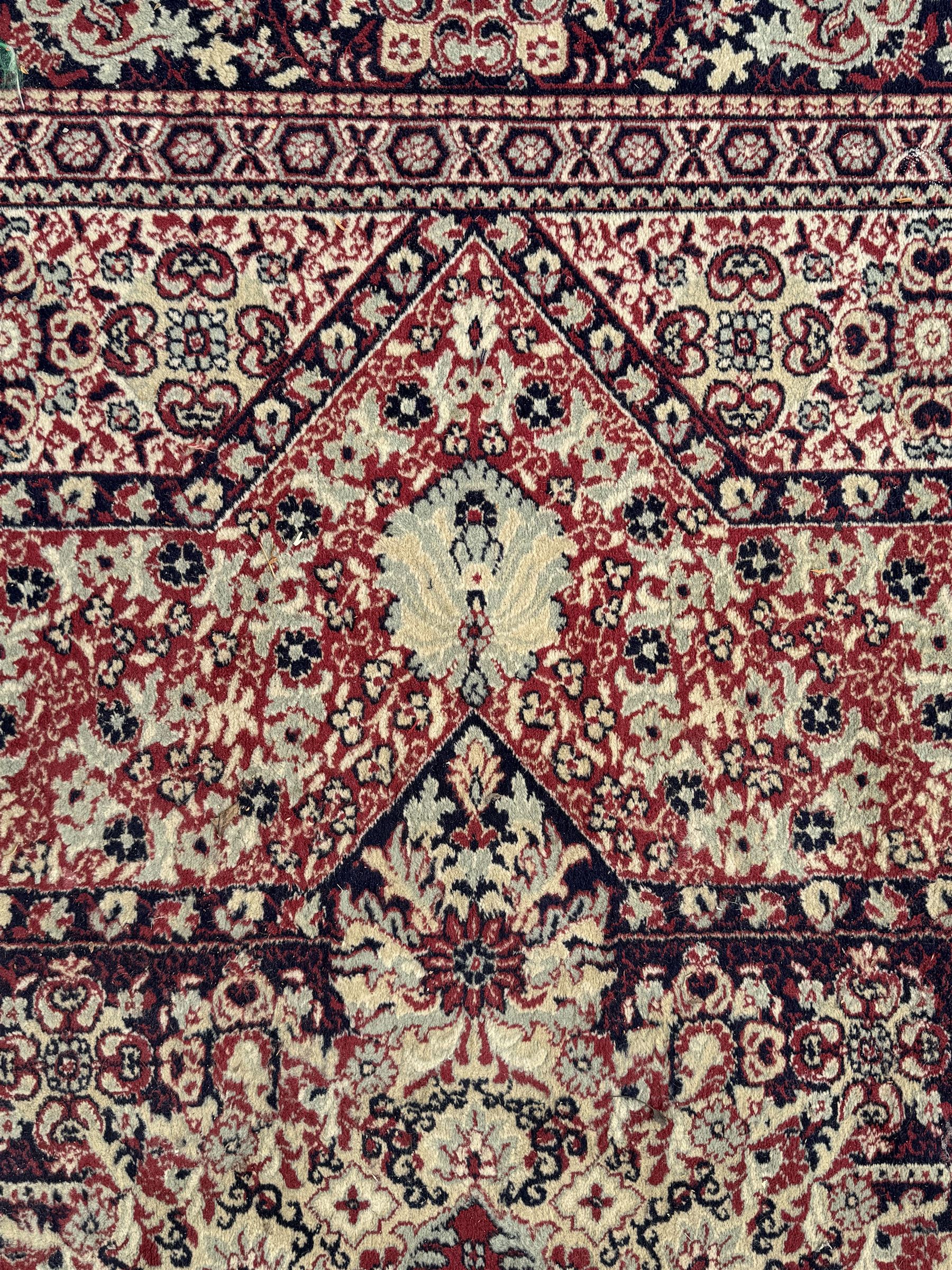 Persian Heriz design red ground carpet, the field decorated with a central star-shaped medallion surrounded by geometric floral motifs and angular vine patterns, the spandrels highlighted with palmette designs, the main border featuring a series of stylised rosettes and leaves against a dark blue ground, enclosed by multiple guard stripes with alternating floral and geometric motifs