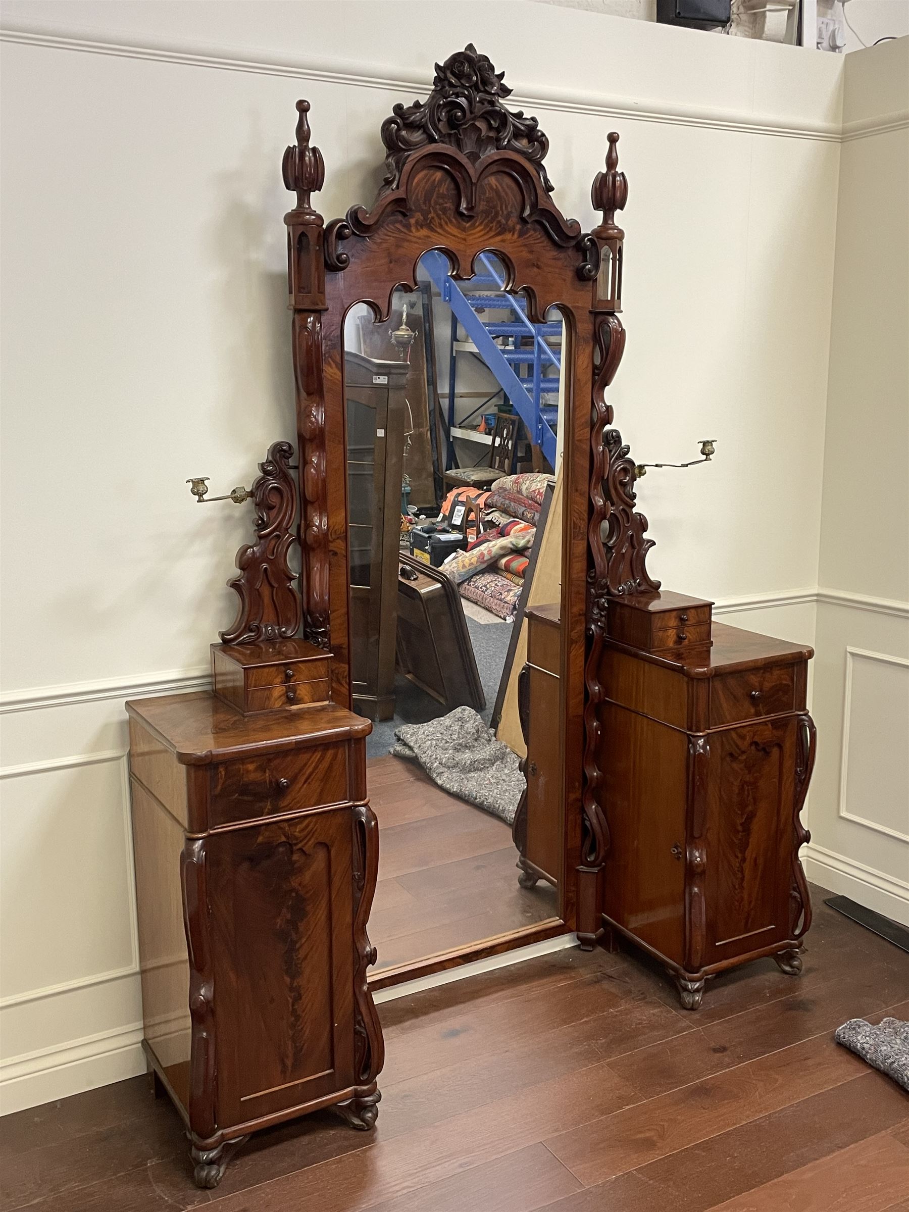 Tall Victorian figured mahogany twin pedestal dressing table, the tall mirror with flower head and scrolled leaf carved pediment, flanked by two turned and carved finials over faceted lancet arched lantern tops, plane mirror plate enclosed by shaped frame, pierced and undulating moulded uprights, on C-scroll and curled leaf carved supports fitted with cast brass hinged candle sconces, each pedestal fitted with two small trinket drawers over larger drawer and panelled cupboard, canted corners with matched upright mounts, the cupboard doors enclosing slides, on scroll carved feet 