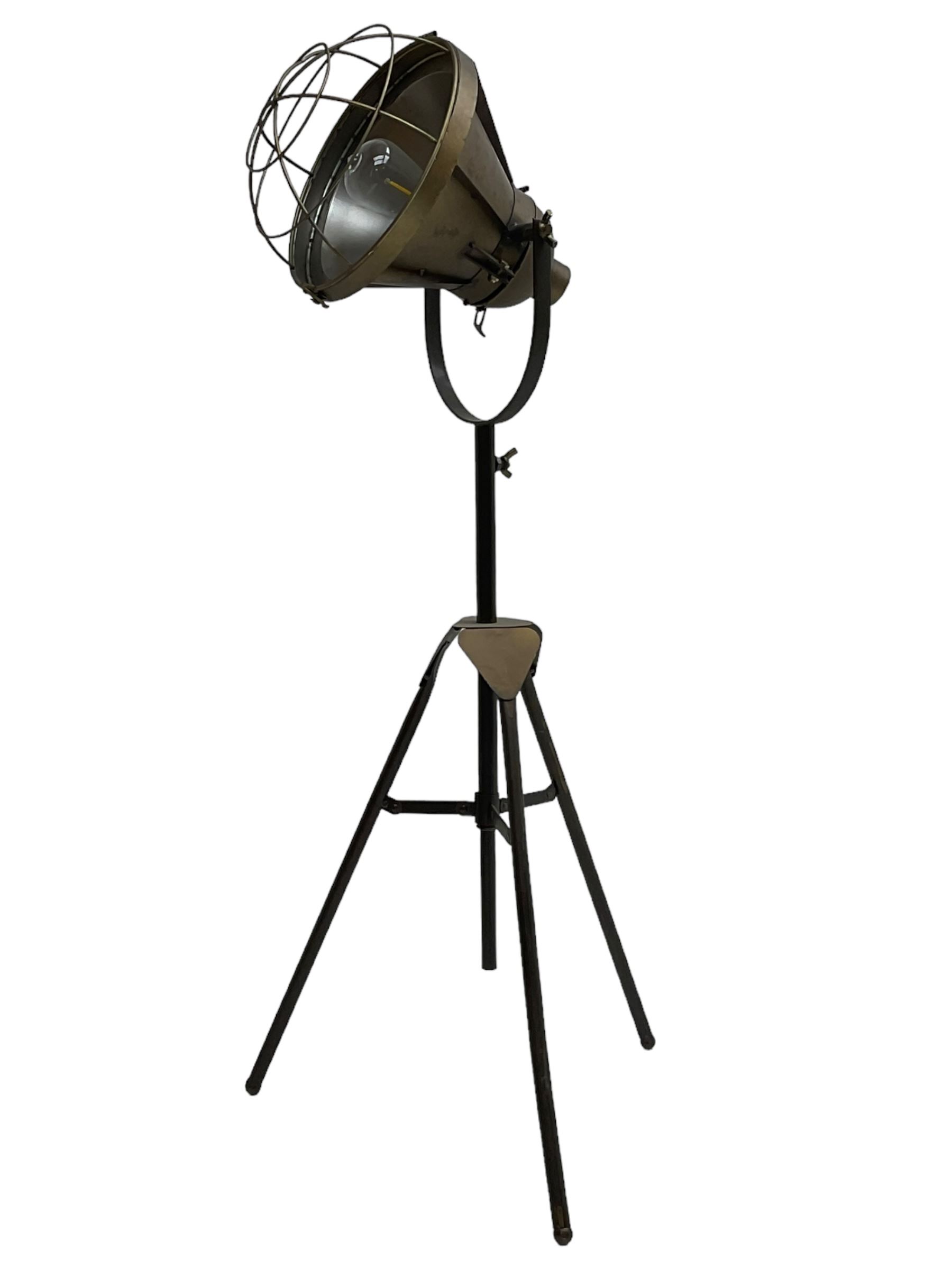 Industrial tripod spotlight, with a metal shade and wire guard, supported by a height-adjustable tripod base, the head equipped with an adjustable tilt mechanism