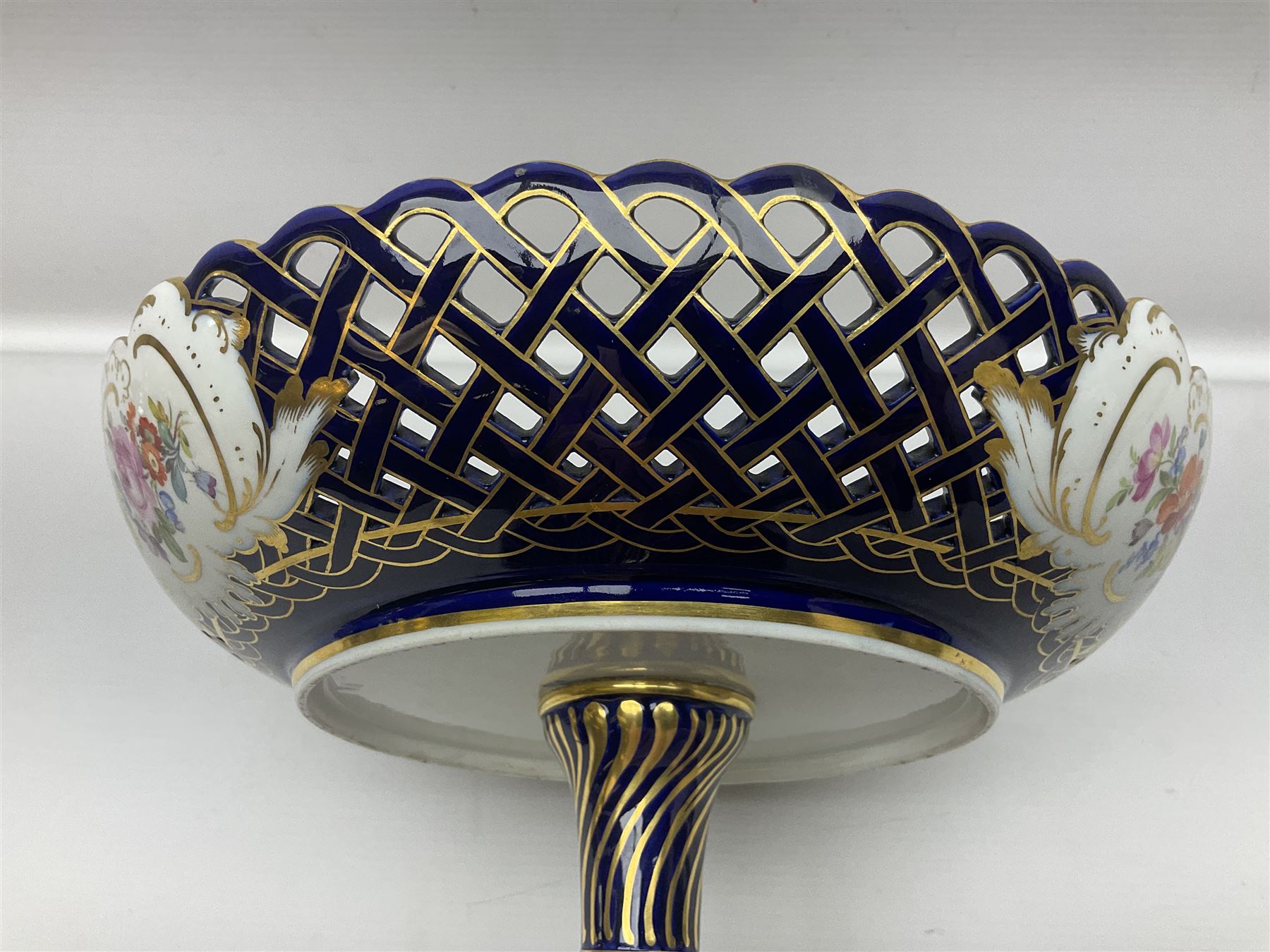 Meissen style pedestal dish, with central panel depicting a trading scene in a harbour setting, the bowl with openwork lattice sides divided by three hand painted floral panels, upon a blue ground with gilt detailing, upon a fluted knopped pedestal and circular foot, H22cm