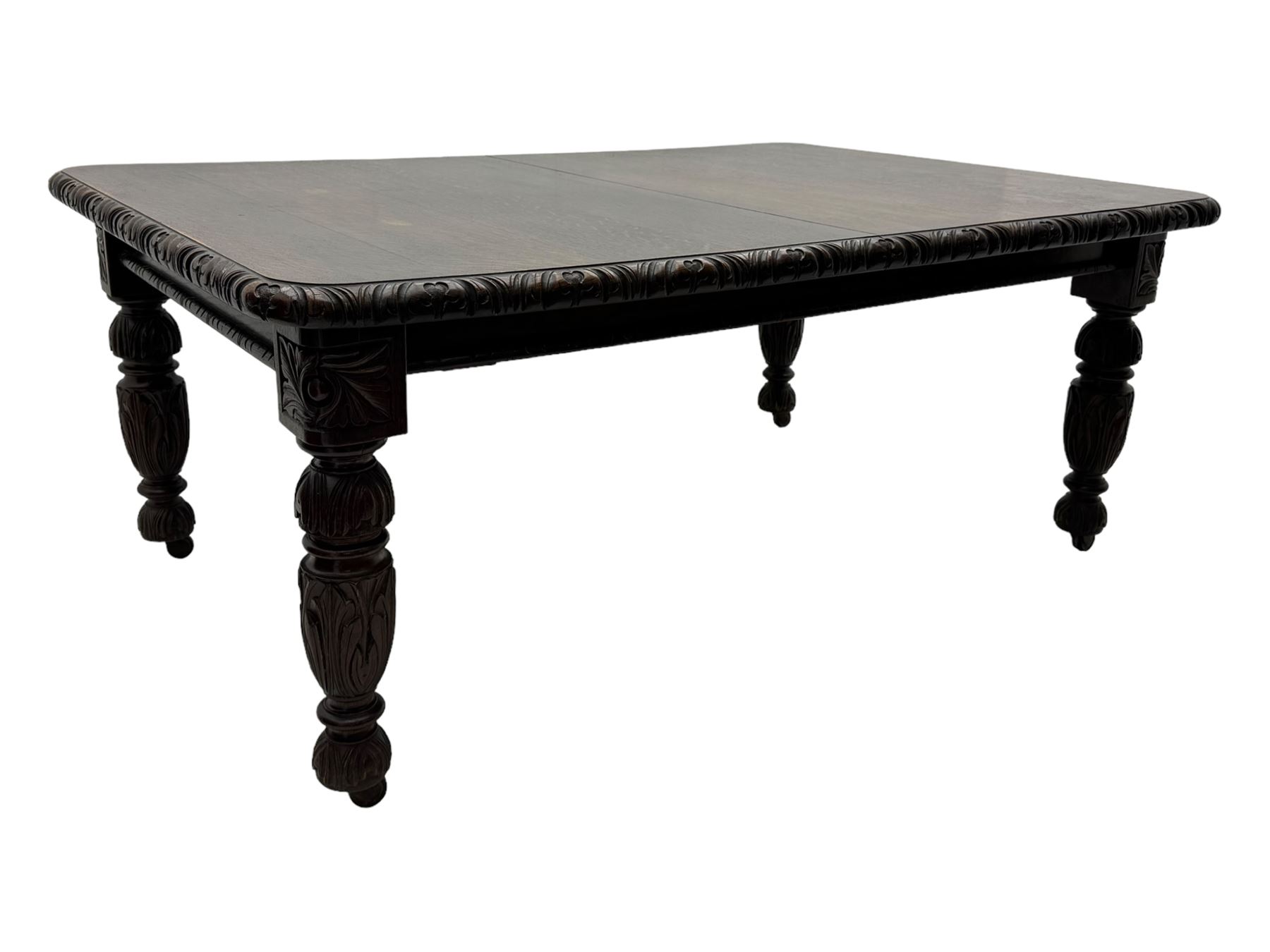 19th century heavily carved oak extending dining table, rectangular top with rounded corners and carved gadrooned edge, extending via winding mechanism with two additional leaves, raised on acanthus leaf-carved baluster supports terminating in ceramic castors