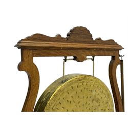 Early 20th century oak gong stand with gong and hammer, shaped cresting rail over moulded upper edge on shaped supports, hanging brass drum gong over fretwork middle rail, on raised platforms terminating to splayed feet