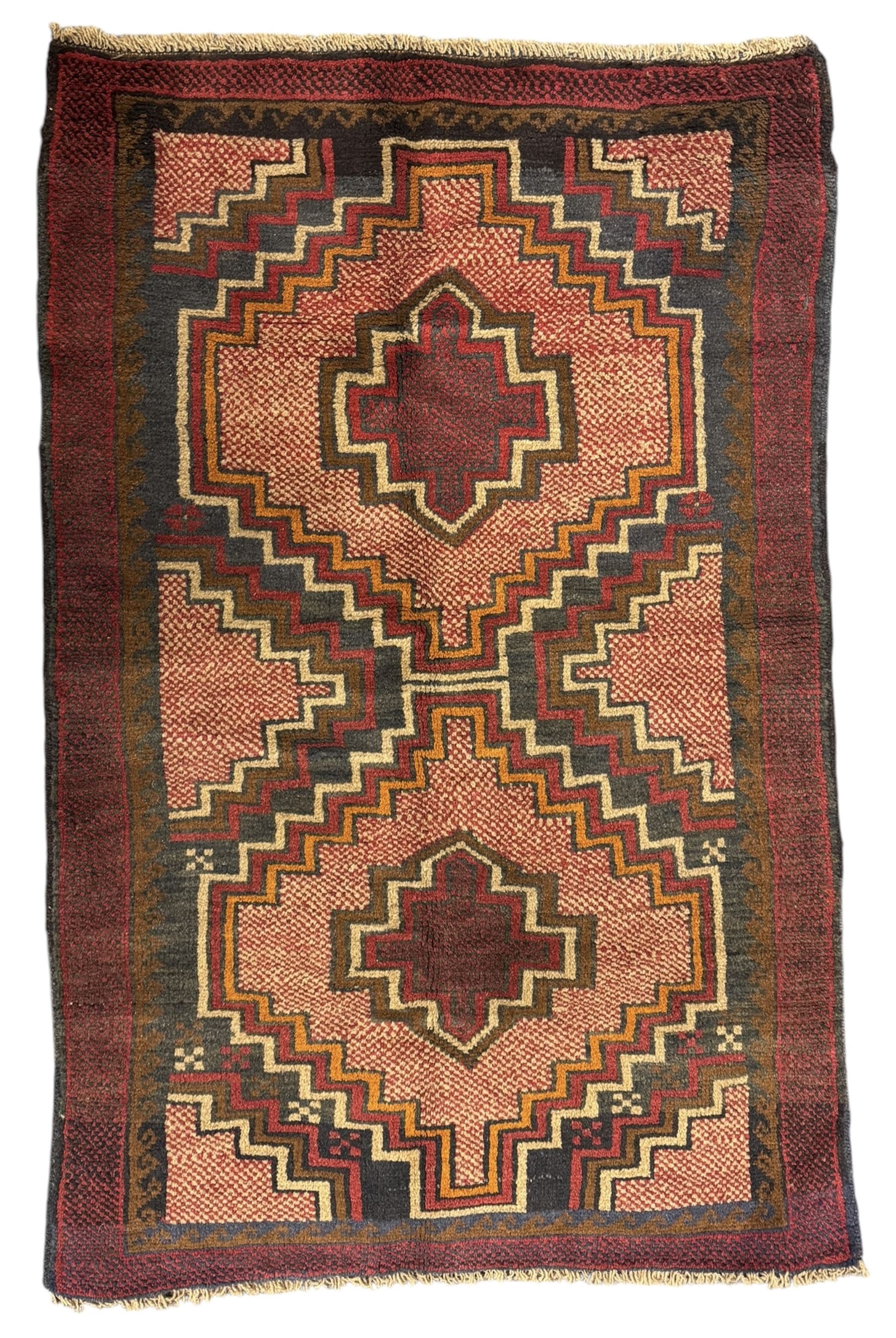 Baluchi crimson ground rug, the central field decorated with two stacked geometric lozenge medallions, surrounded by stepped multicoloured motifs, the wide border with alternating angular motifs in deep red, within a narrow brown guard stripe