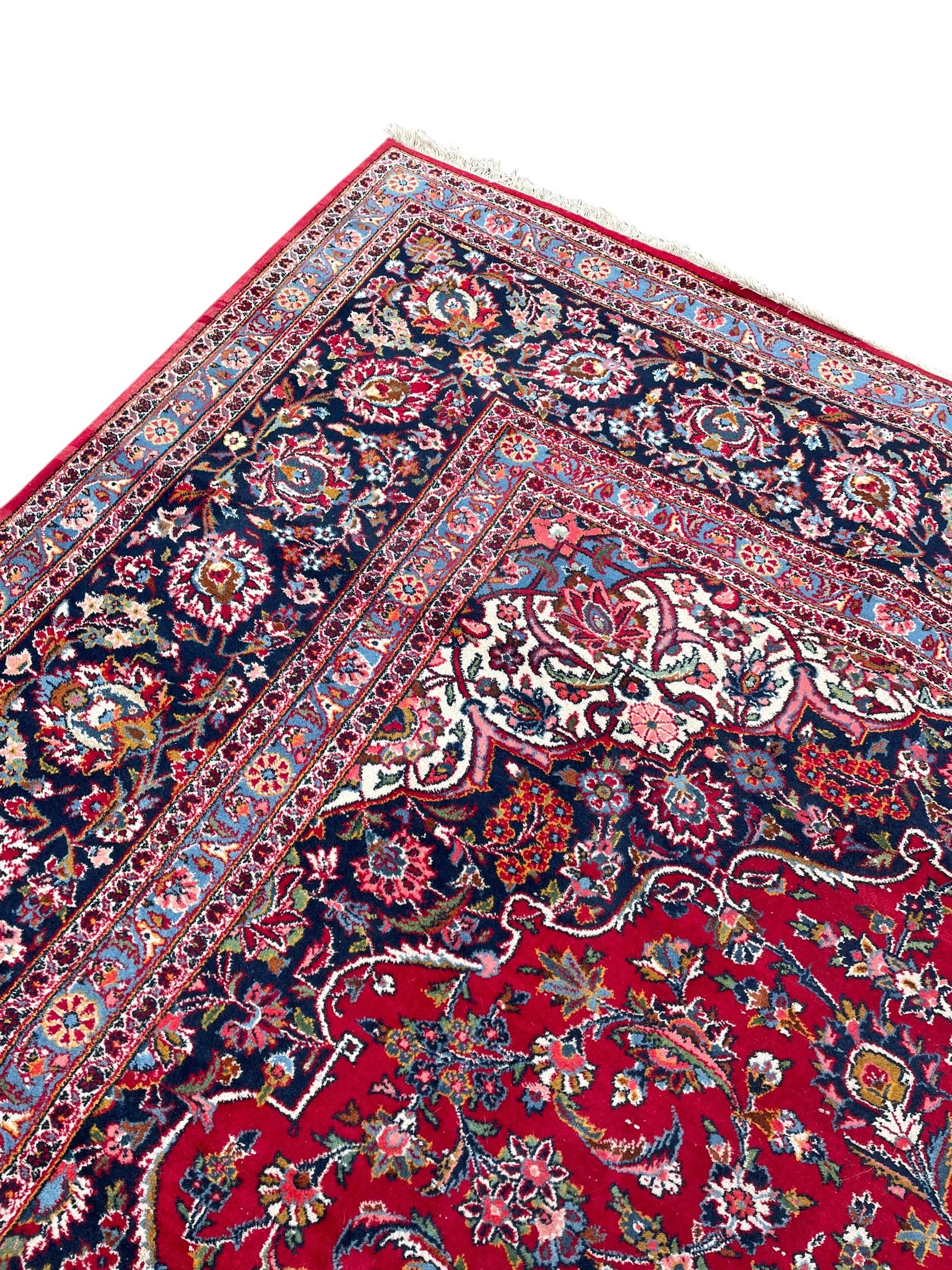 Large Persian Kashan crimson ground carpet, central floral medallion surrounded by swirling leafy branches and palmettes, enclose by floral pattern spandrels, the indigo border with overall scrolling design decorated with palmettes, within guard stripes 