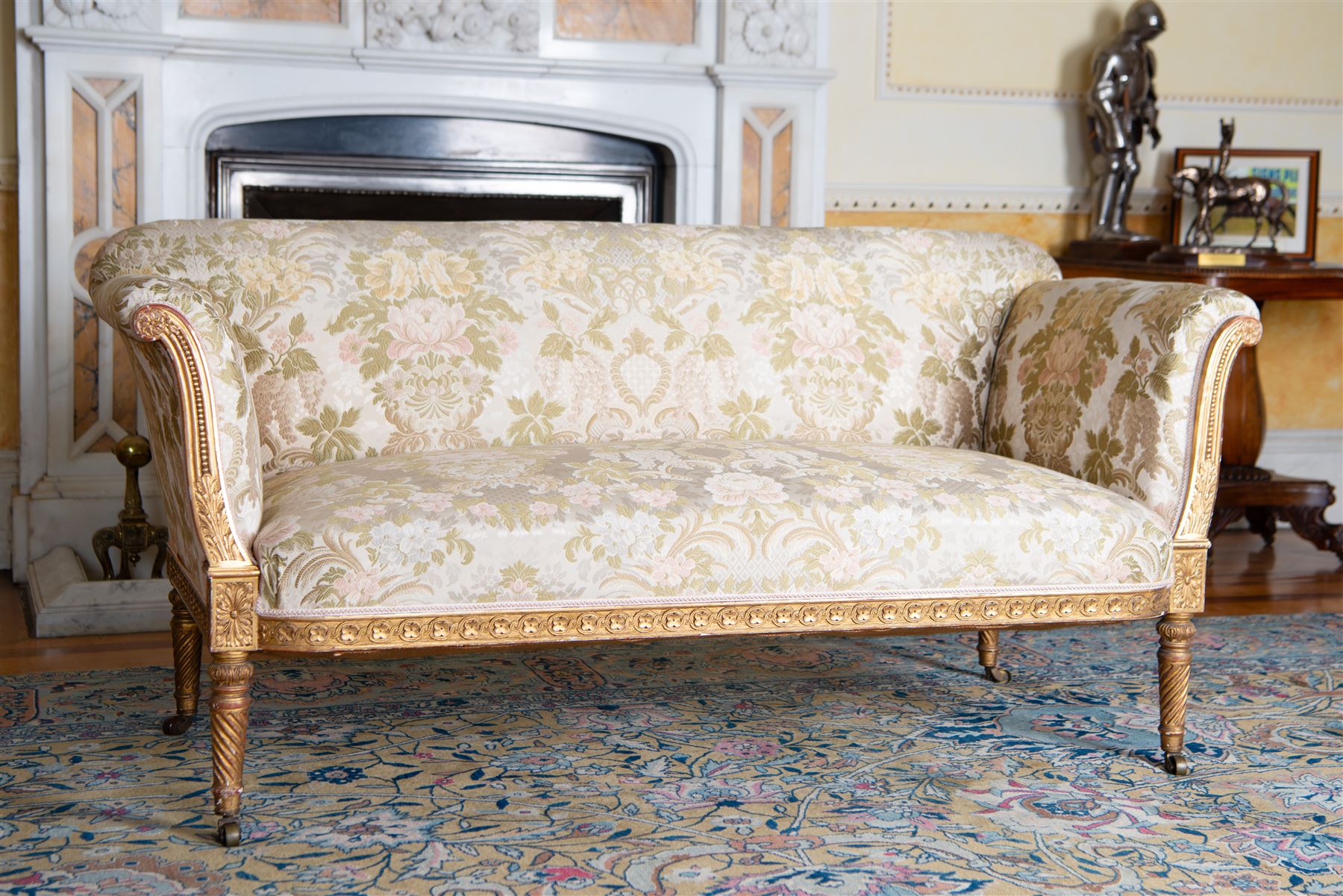 19th century gilt wood and gesso settee, upholstered in repeating floral pattern fabric, rolled arms decorated with trailing bead moulding and acanthus leaf, the seat rail with flower head band, on twist turned and foliate carved feet with brass castors 