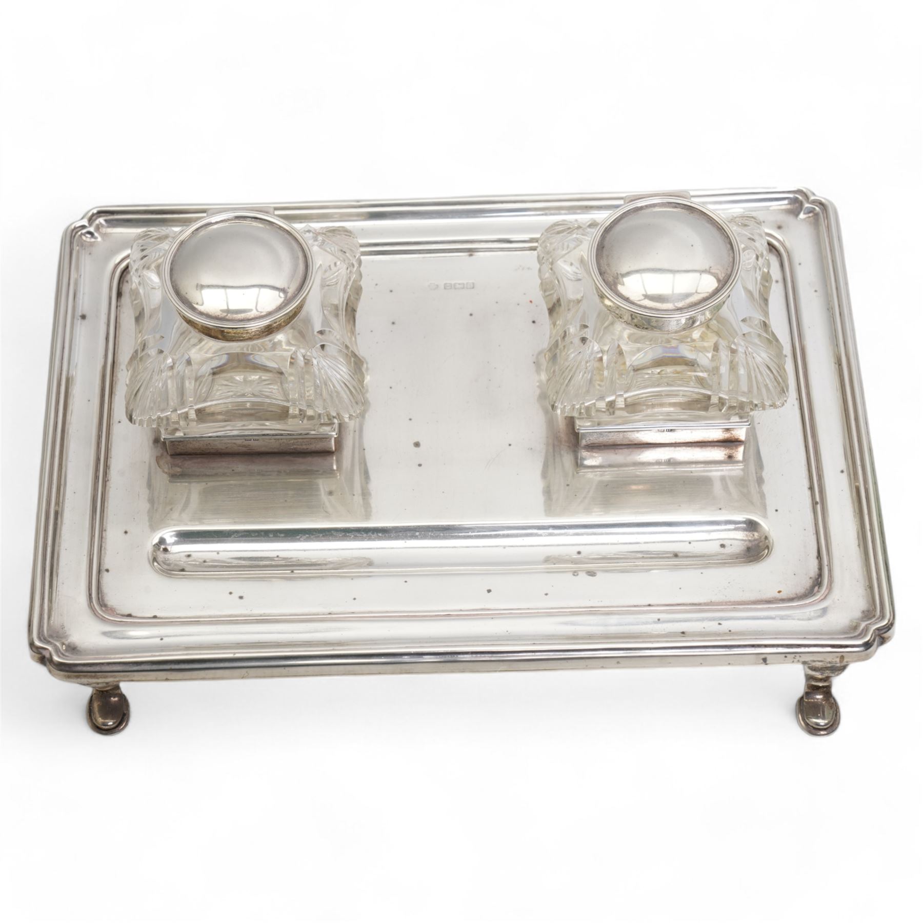 George V silver inkstand of rectangular design with pen tray and two cushion shape glass inkwells with silver covers, raised edge and four shaped supports 28cm x 20cm Birmingham 1912 Maker William Hutton & Sons