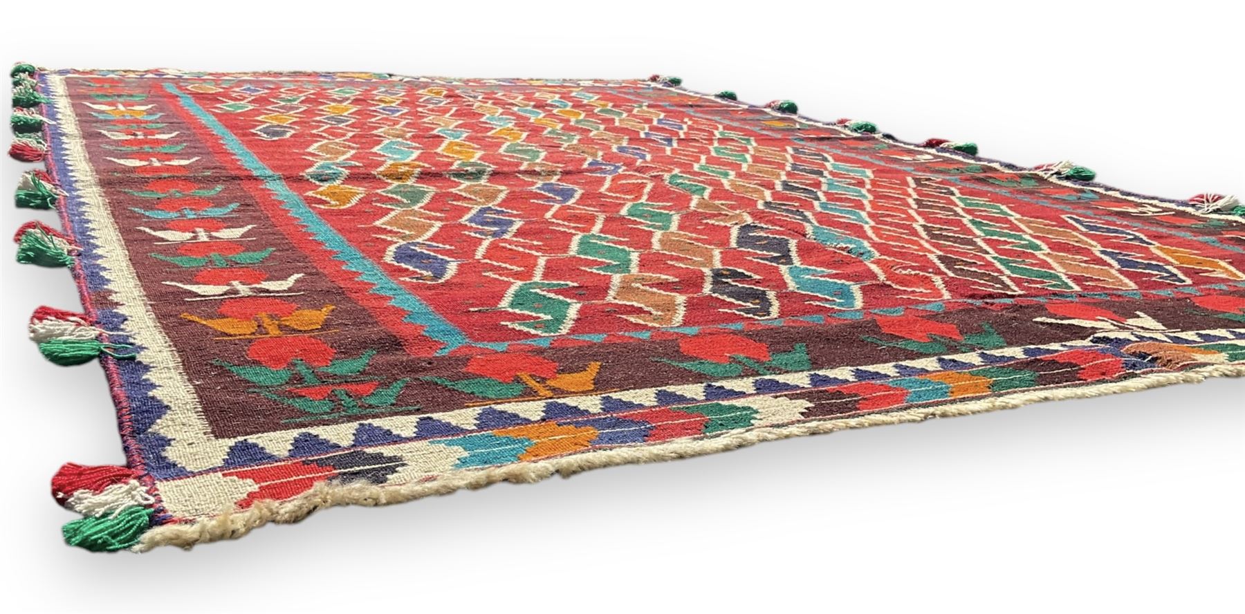 Southwest Persian Qashqai Kilim crimson ground rug, decorated with a repeating geometric pattern of interlocking diamond motifs in red, green, blue, and brown, enclosed by a deep brown border with stylised floral motifs and colourful fringe accents