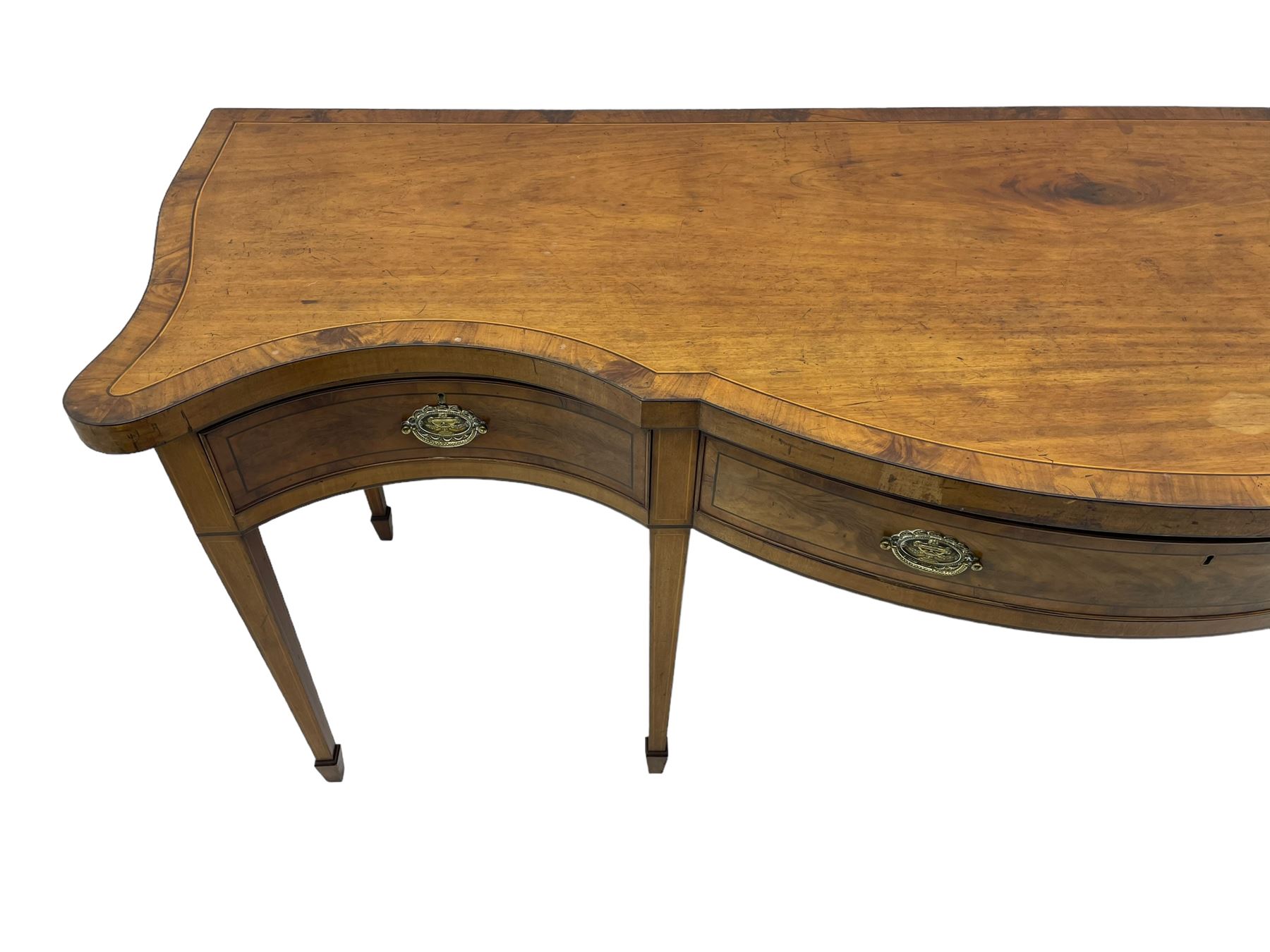 Large George III mahogany serpentine serving table, shaped top with crossing banding and stringing, the frieze fitted with three cock-beaded drawers, oval pressed brass handle plates decorated with urns, on square tapering supports with spade feet 