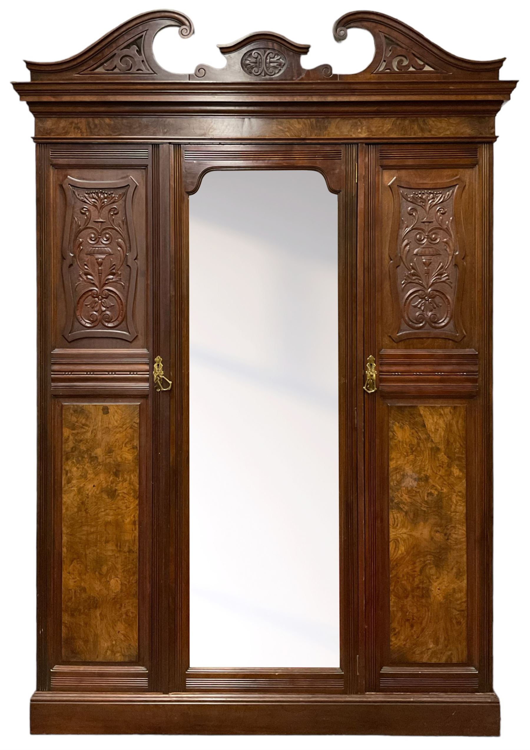 Late Victorian walnut triple wardrobe, the pierced broken swan neck pediment carved with foliate C-scrolls, central bevelled mirror door flanked by two panelled doors, the panels carved with urns, C-scrolls and trailing foliage, the interior fitted with hanging rails, long and short drawer, on moulded plinth base