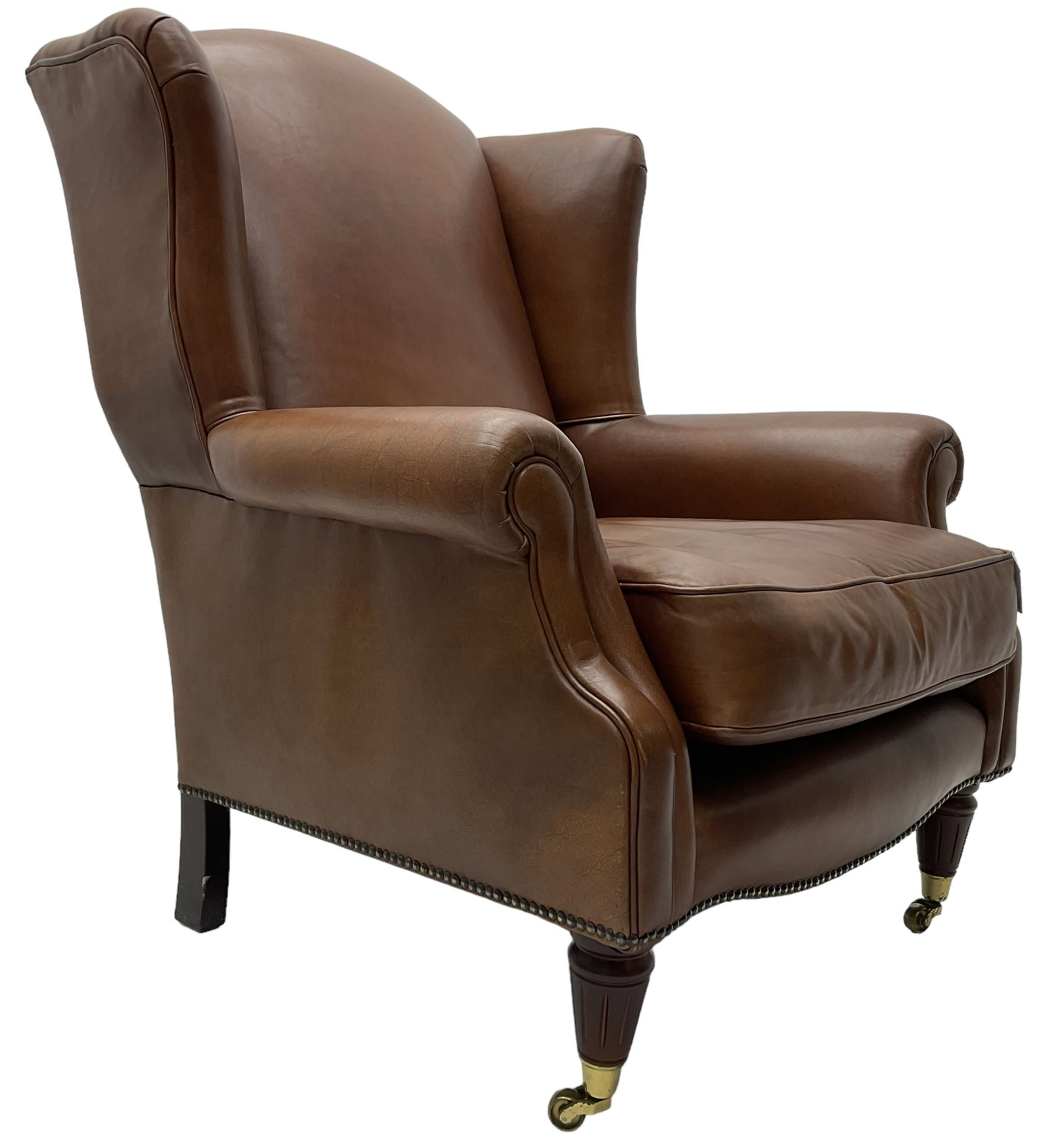 Georgian design wingback armchair, upholstered in brown leather with stud band and piping, turned and fluted front feet on brass cups and castors