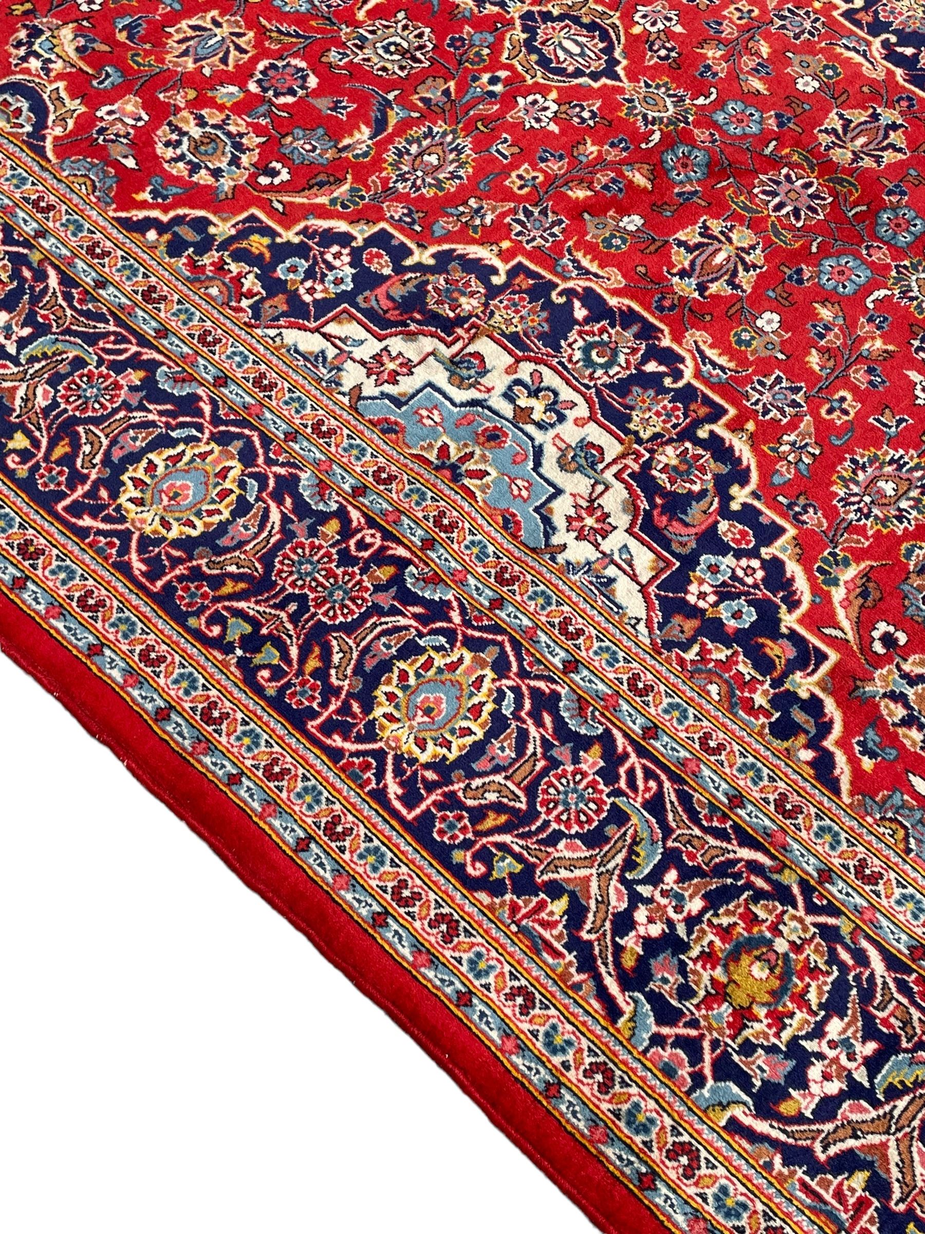 Persian Kashan crimson ground rug, overall arabesque design, the field decorated with swirled leafy branches and palmettes, indigo ground border decorated with repeating pattern, within guard stripes 