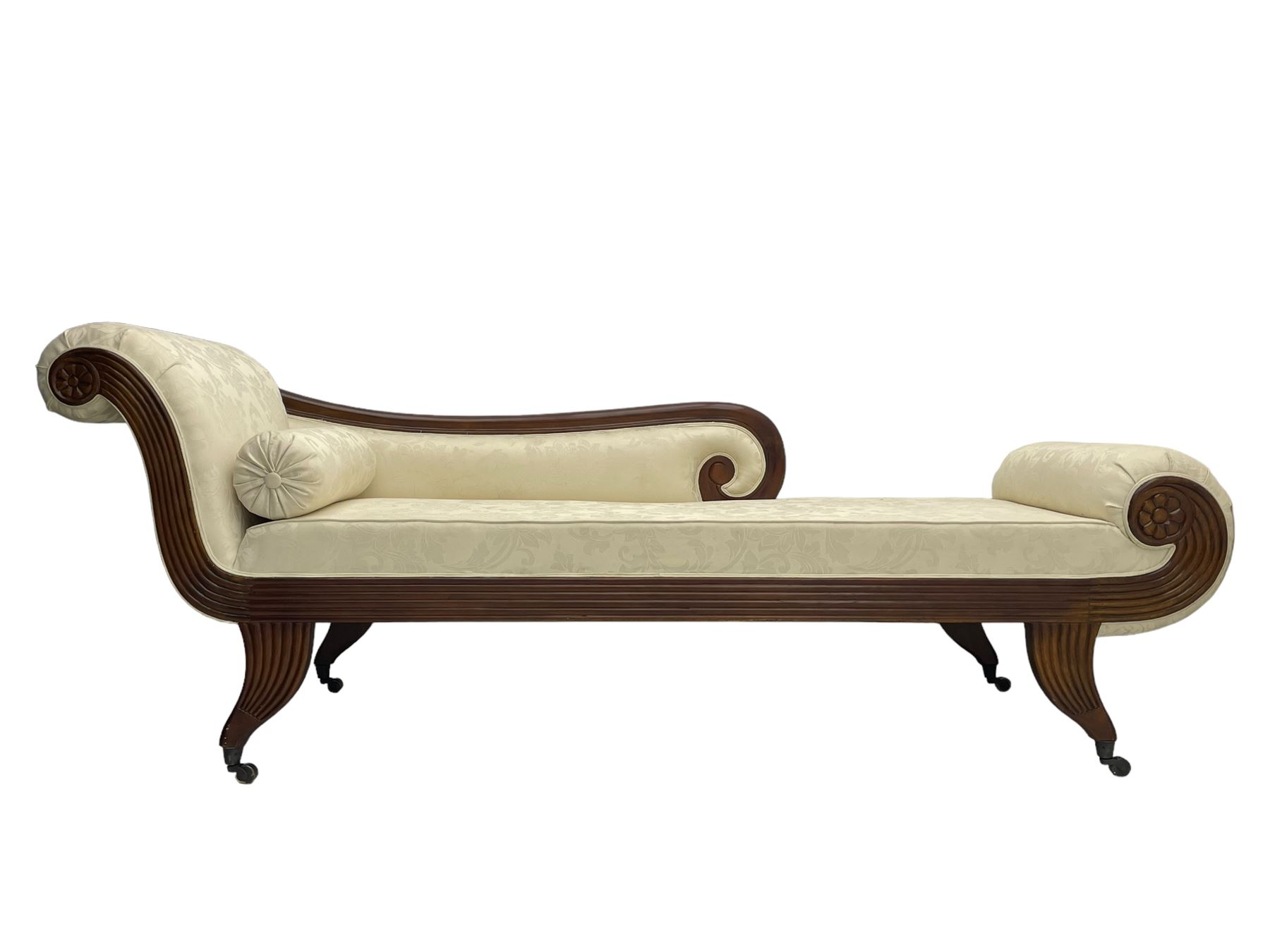 Regency design mahogany chaise longue, scrolled arms with carved rosette details, upholstered in cream damask fabric with bolster cushion, reeded frame supported by turned legs on brass castors