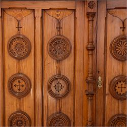 19th century pitch pine ecclesiastical vestment wardrobe or sacristy cabinet, crenelated and projecting moulded cornice over two panelled doors, the panelled doors carved with linen folds and mounted with rows of roundels depicting crucifixes and flower heads, enclosed by turned pilasters, two drawers to the base, on moulded plinth base 