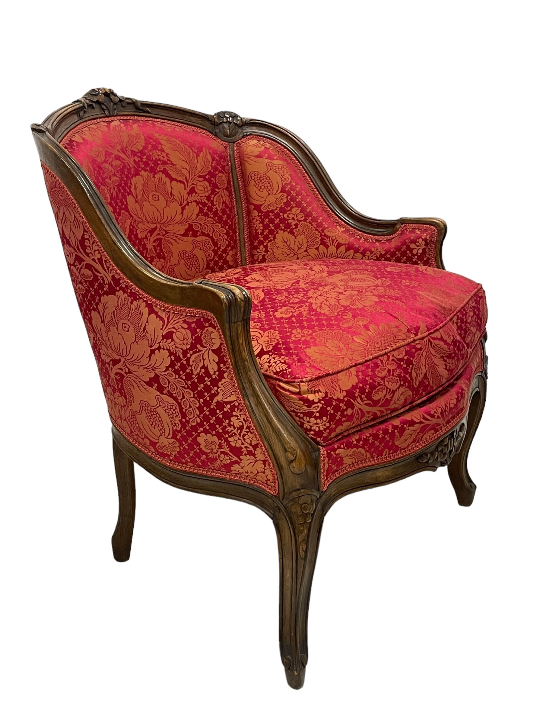 Late 20th century French design stained beech framed armchair and matching stool - the armchair with shaped and moulded frame carved with flower heads and foliage, upholstered in silk red and gold ground fabric with floral pattern, on scroll carved cabriole feet decorated with flower head carvings (W73cm, H82cm, D75cm); the footstool of concaved rectangular form with moulded frame carved with flower heads, upholstered loose cushion, on cabriole feet (70cm x 64cm, H48cm)