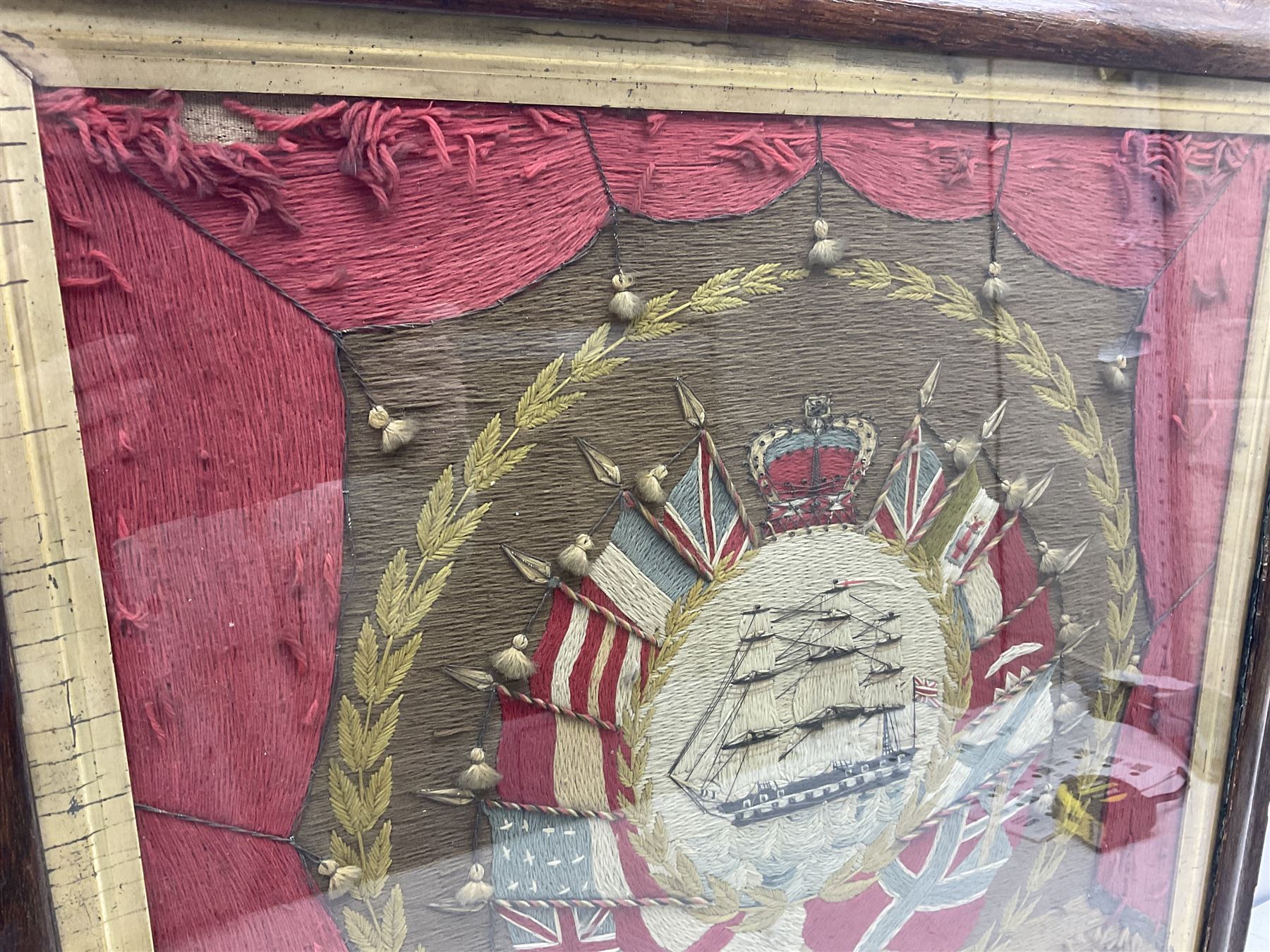 19th century woolwork, depicting a clipper ship, fully rigged over black hull with white ports, within a border of ensigns and flags and beaded crown above, in glazed wooden frame, H57cm