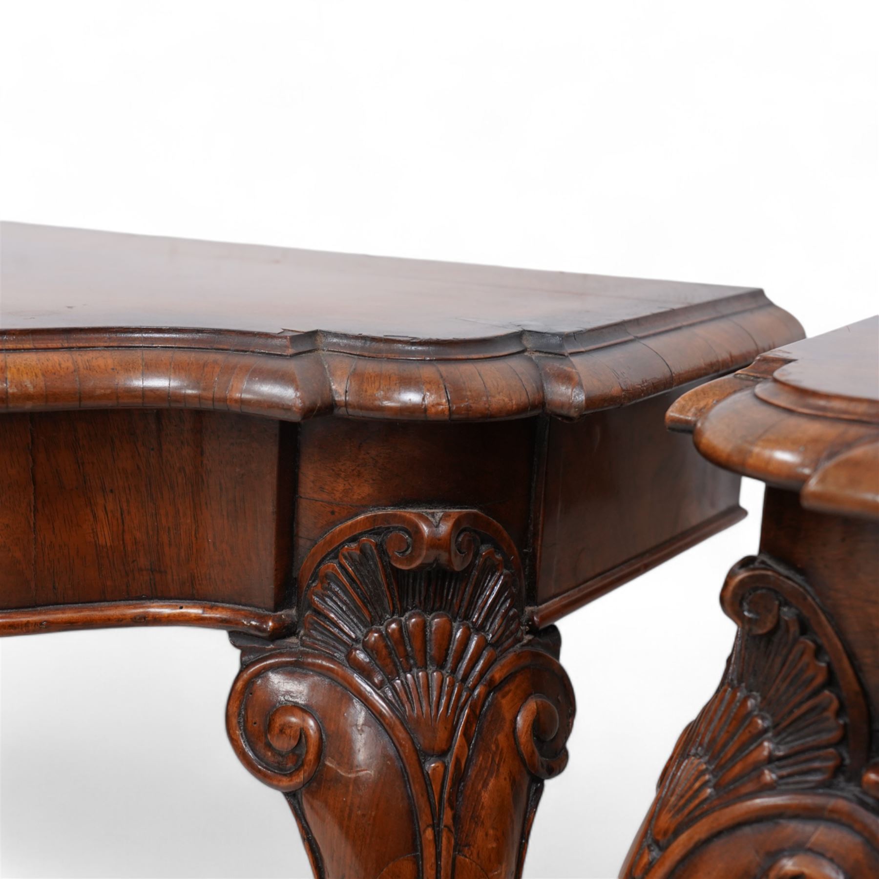 Pair of Georgian walnut console tables, the shaped rectangular cross-banded top with moulded edge over a banded frieze, raised on moulded cabriole supports decorated with scalloped shell and scroll carved knees, terminating to hairy hoof feet