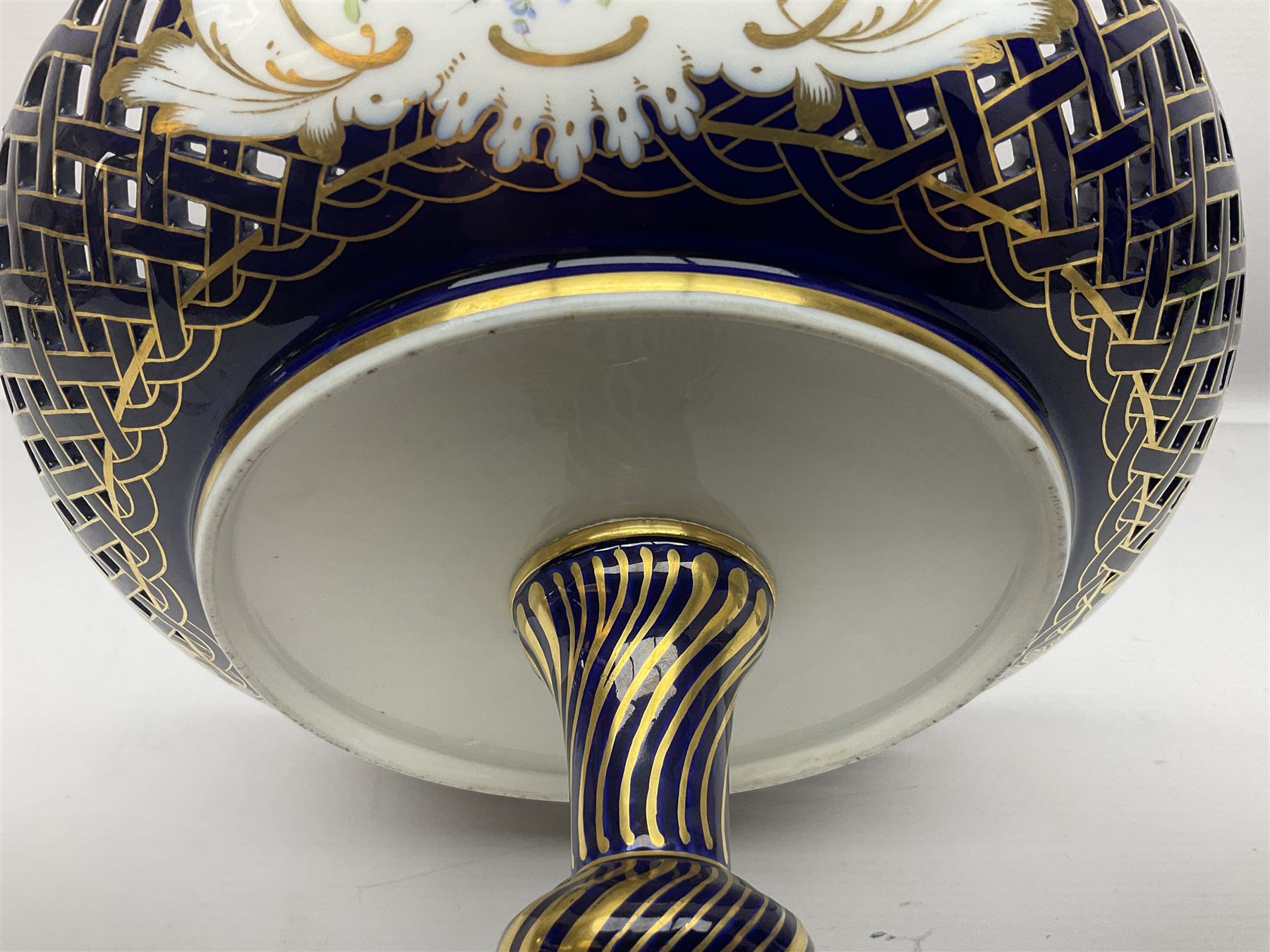 Meissen style pedestal dish, with central panel depicting a trading scene in a harbour setting, the bowl with openwork lattice sides divided by three hand painted floral panels, upon a blue ground with gilt detailing, upon a fluted knopped pedestal and circular foot, H22cm