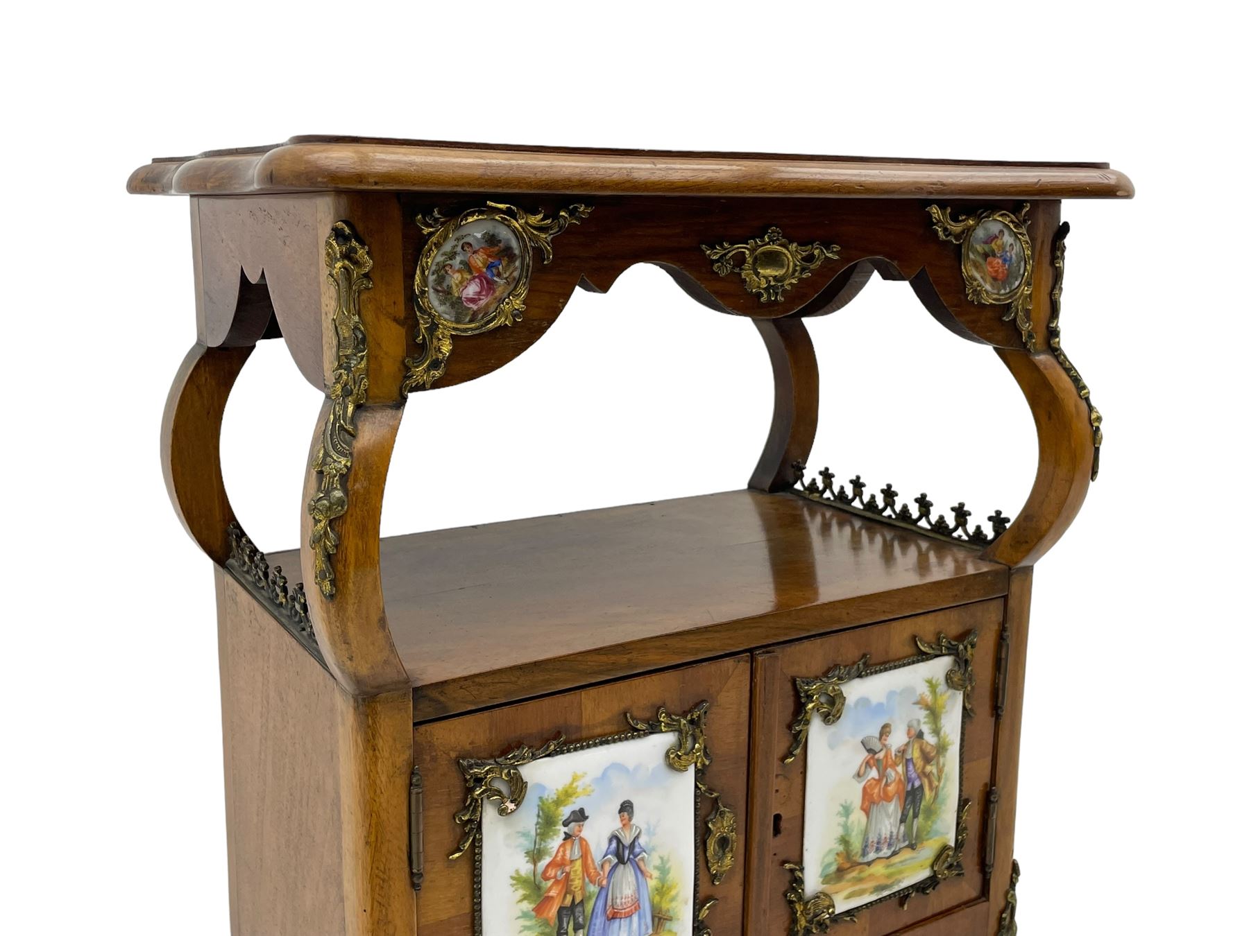 Mid-to-late 20th century French figured walnut three-tier bedside stand, shaped moulded top inlaid with figured book-matched veneers and crossbanding, double cupboard below enclosed by two doors mounted by painted porcelain panels depicting courting scenes within landscapes, on cabriole supports united by undertier, decorated with ornate cast metal cartouche mounts 