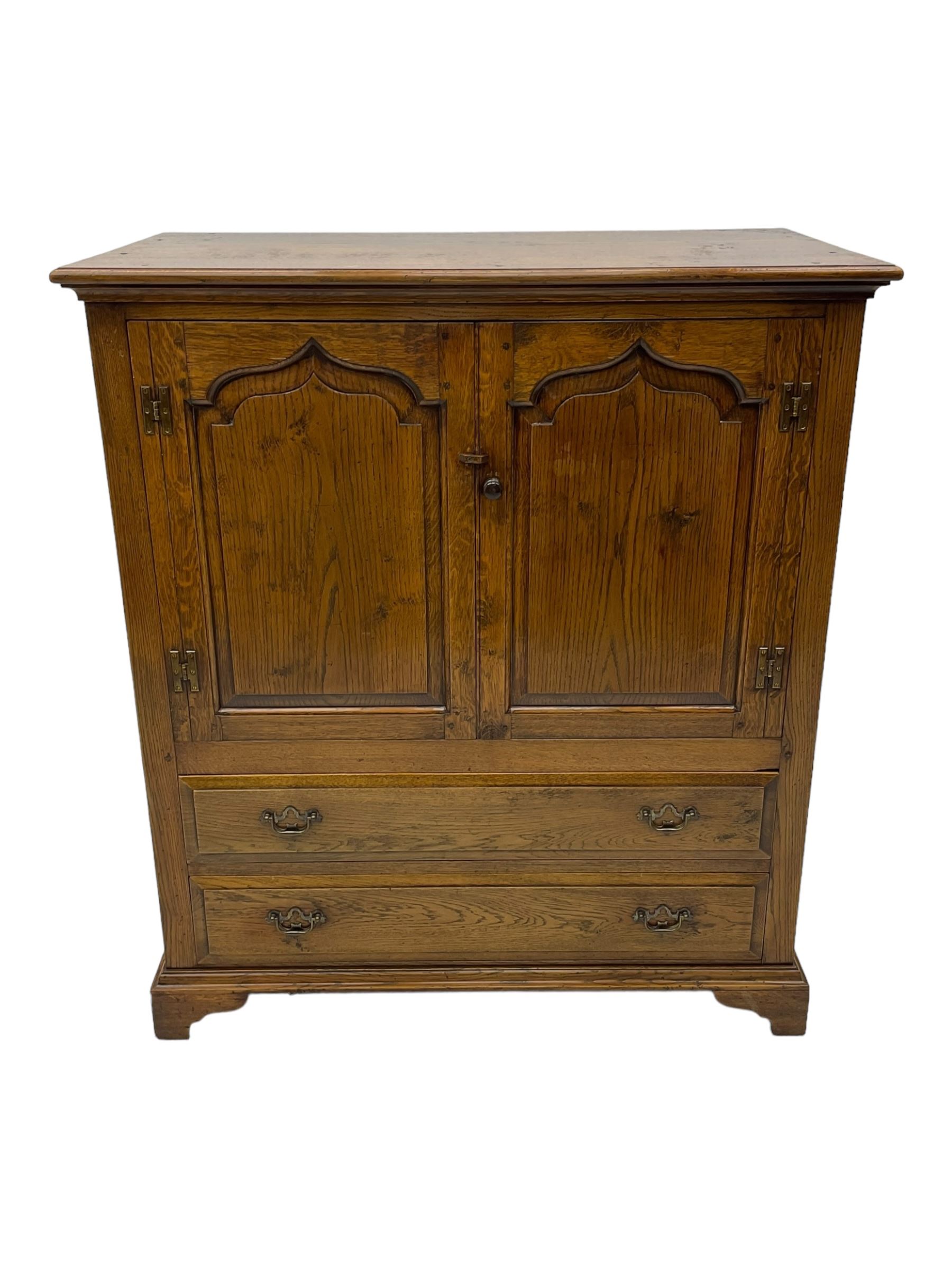 Oak drinks or media cabinet, projecting moulded cornice over two Gothic arch panelled doors on sliding tracks enclosing open storage, two drawers with brass drop handles, on bracket supports