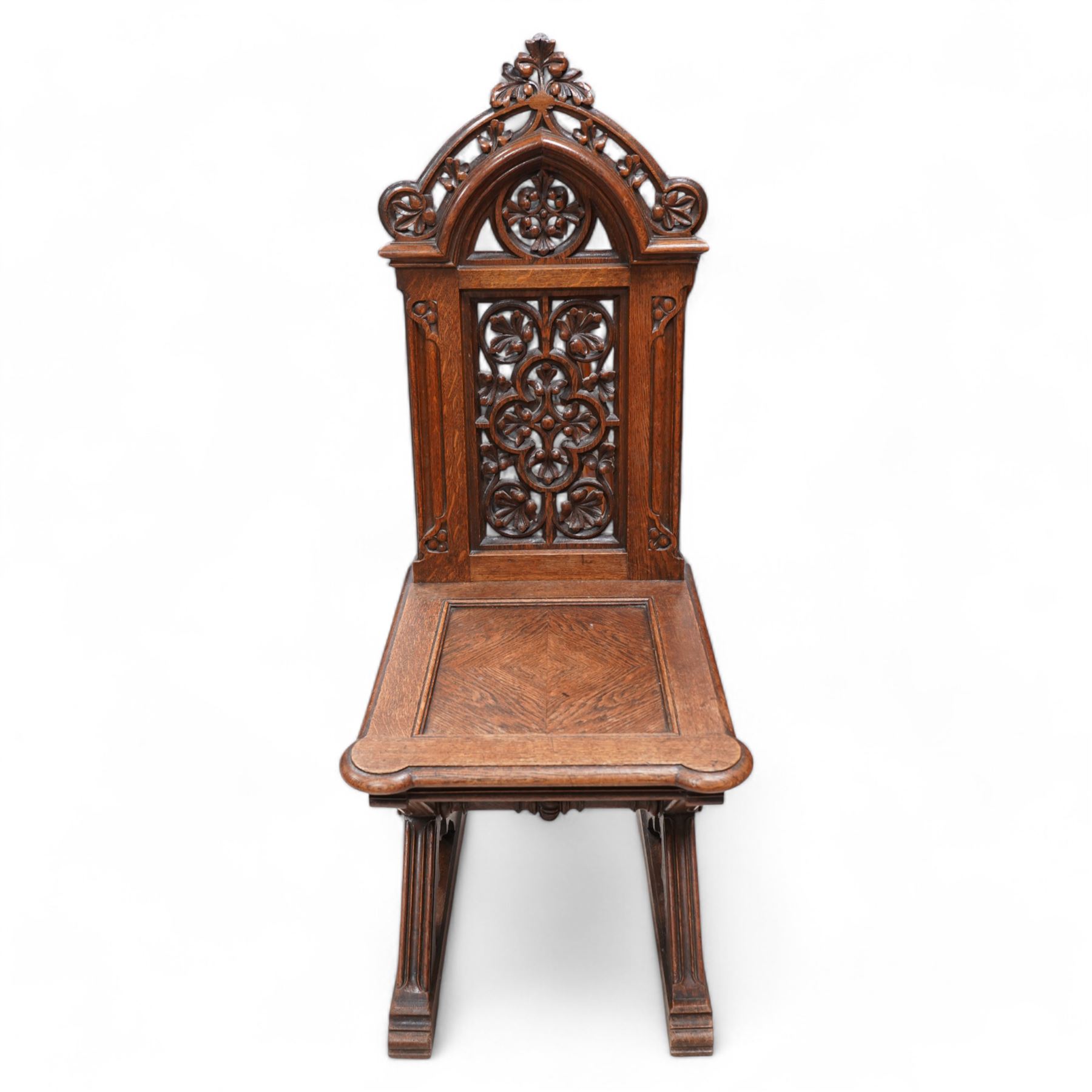 In the manner of Augustus Pugin - 19th century Gothic Revival carved oak hall chair, the pierced and carved arched pediment pierced and carved with scrolling foliage, the pierced back panel carved with a central pierced quatrefoil and leafage, panelled and eared seat over an architectural X-frame base with chamfered trefoil carvings and sledge feet