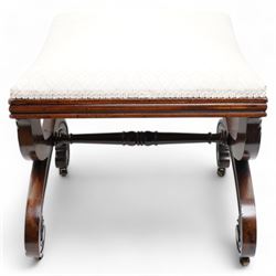 Early 19th century mahogany stool, the dished seat upholstered in cream fabric with repeating pattern, moulded seat rails, on curved X-frame supports carved with scrolls and foliage, united by turned stretcher, on brass castors