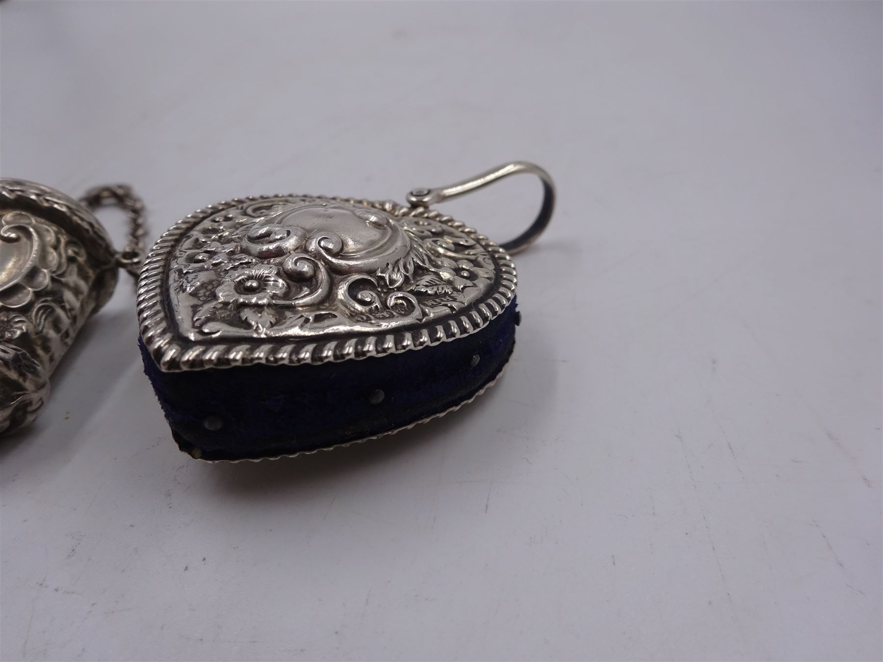 Edwardian silver pin cushion, in the form of a heart, with embossed floral decoration, with blue pin cushion between two heart shaped silver covers, with chatelain hook, hallmarked Henry Matthews, Birmingham 1902, together with a late Victorian silver thimble holder, embossed with scrolling decoration and with velvet interior, with chatelain chain, hallmarked Levi & Salaman, Birmingham 1899, pin cushion H4.5cm