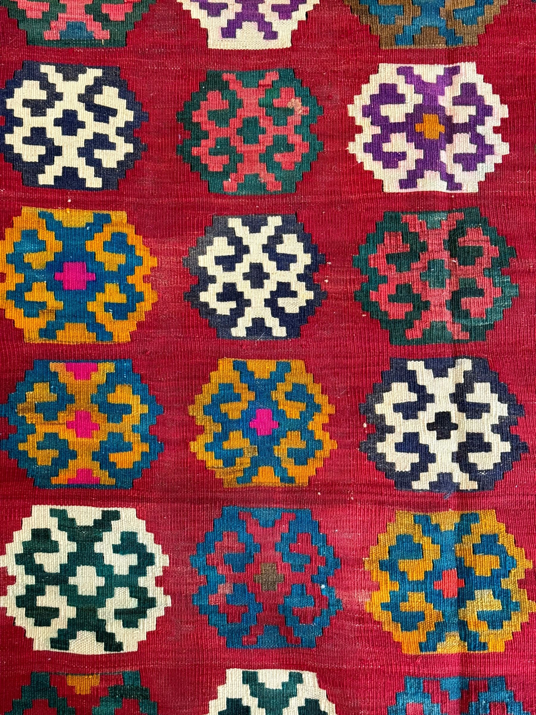 Southwest Persian Qashqai Kilim red ground rug, the central field adorned with rows of multicolored hexagonal medallions with geometric patterns, the border featuring alternating black and white stepped diamonds, narrow band to one end decorated with multicoloured geometric figures